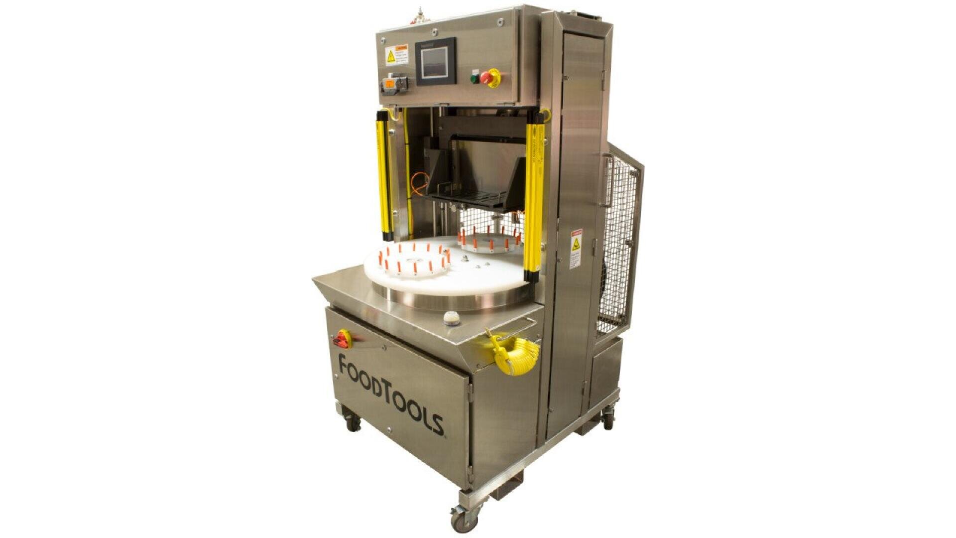 Eine industrielle Lebensmittelverarbeitungsmaschine aus rostfreiem Stahl mit einem Bedienfeld, gelben Schutzvorrichtungen und einem rotierenden weißen runden Tablett mit der Aufschrift FOODTOOLS auf der Vorderseite, das auf Rollen steht.
