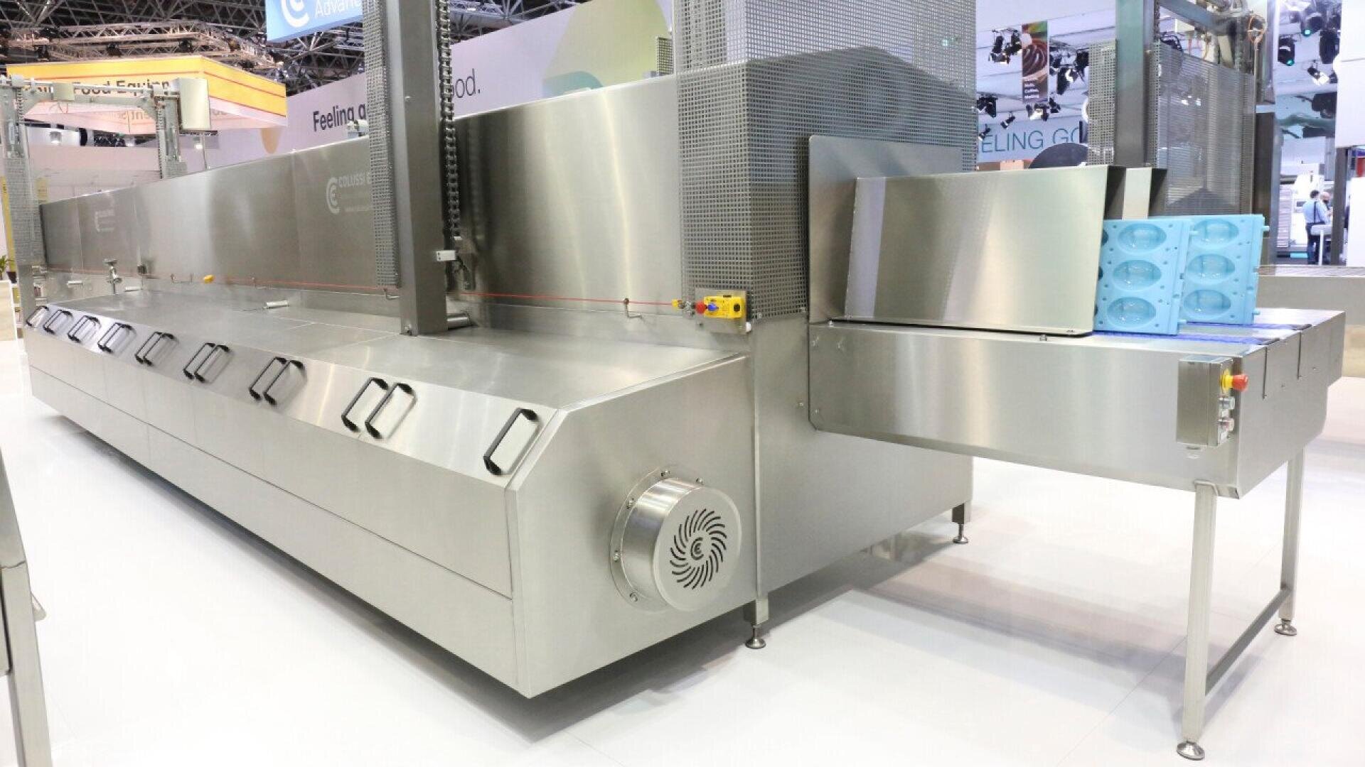 A large, industrial stainless steel food processing machine with vented panels and a conveyor belt, displaying blue plastic trays with molds at the end. The machine is set up in an exhibition setting.