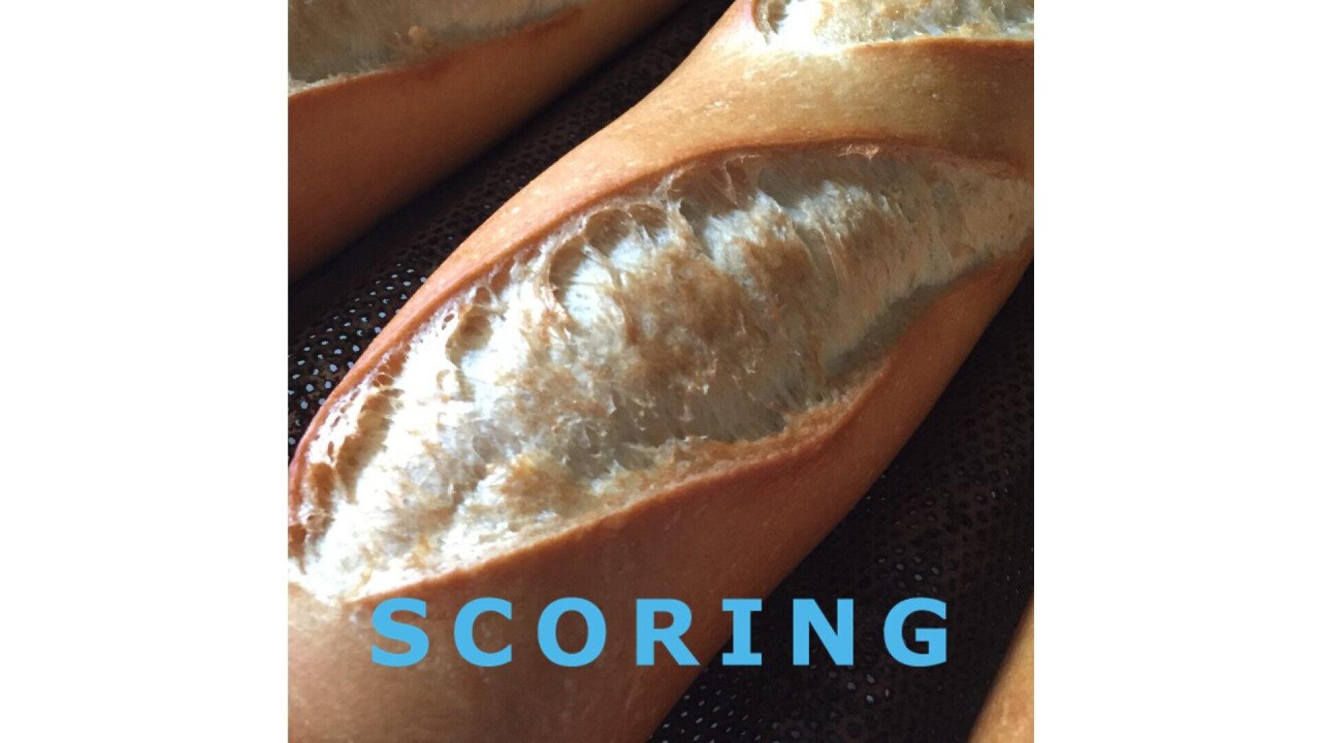 Eine Nahaufnahme eines frisch gebackenen Baguettes mit einem tiefen, gut definierten Einschnitt auf der Oberseite, der die Technik des Einkerbens von Brot zeigt. Das Wort SCORING erscheint in blauer Schrift am unteren Rand des Bildes.