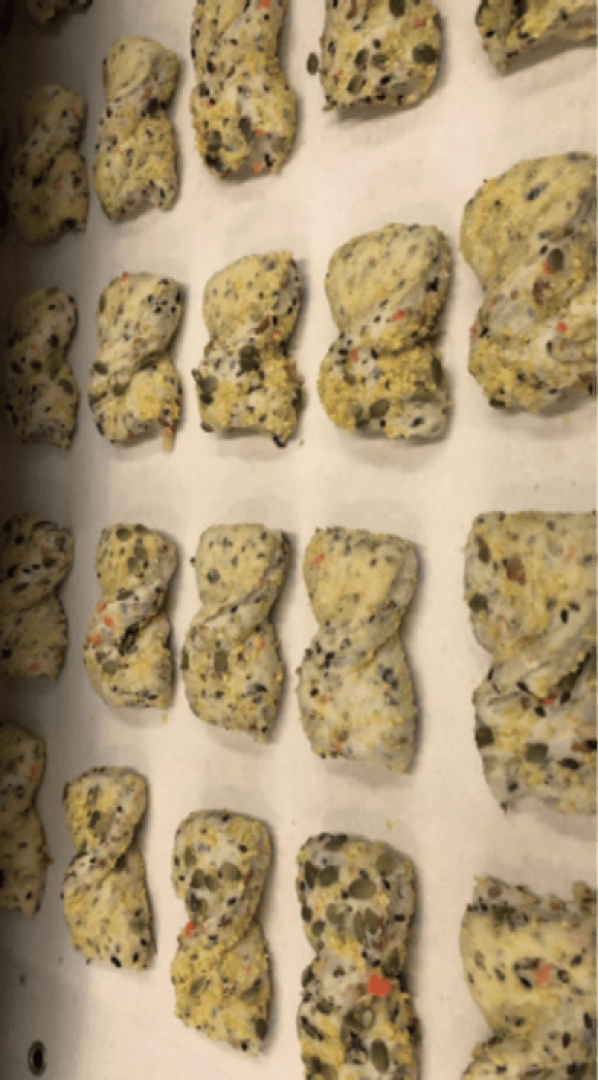 Rows of uncooked, rectangular dough pieces filled with seeds and herbs are arranged on a parchment-lined baking sheet, ready to be baked.