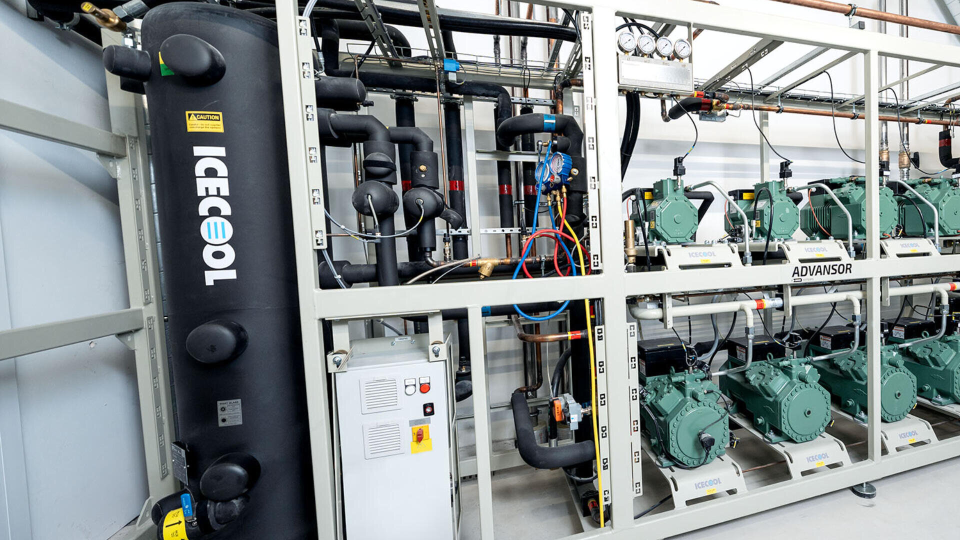 Ein kommerzielles Kühlsystem mit mehreren grünen Kompressoren, beschrifteten Komponenten, Messgeräten, Rohren, Kabeln und einem großen schwarzen vertikalen Tank mit der Aufschrift ICECOOL in einer Industrieanlage.
