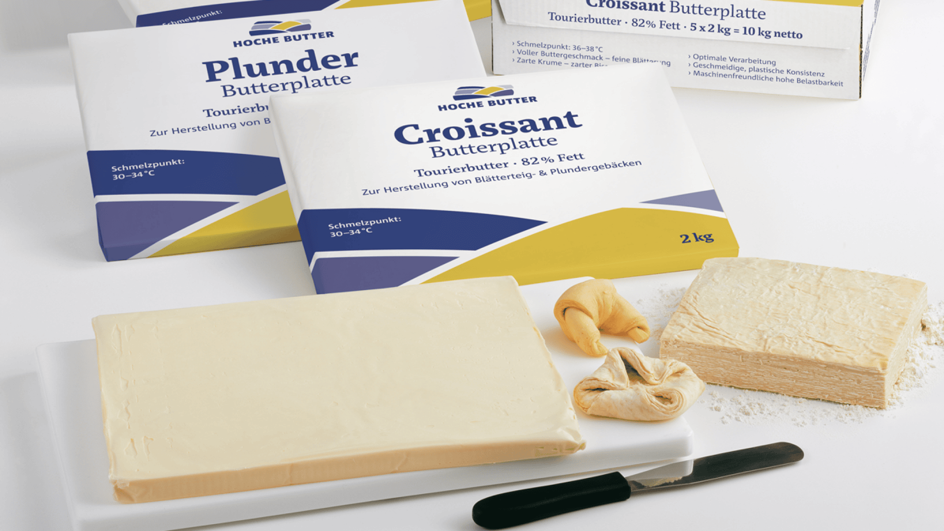 Rechteckige Butterblöcke auf einem weißen Tablett mit einem Messer, zwei kleinen Croissants und drei Verpackungen mit der Aufschrift Plunder Butterplatte und Croissant Butterplatte im Hintergrund.