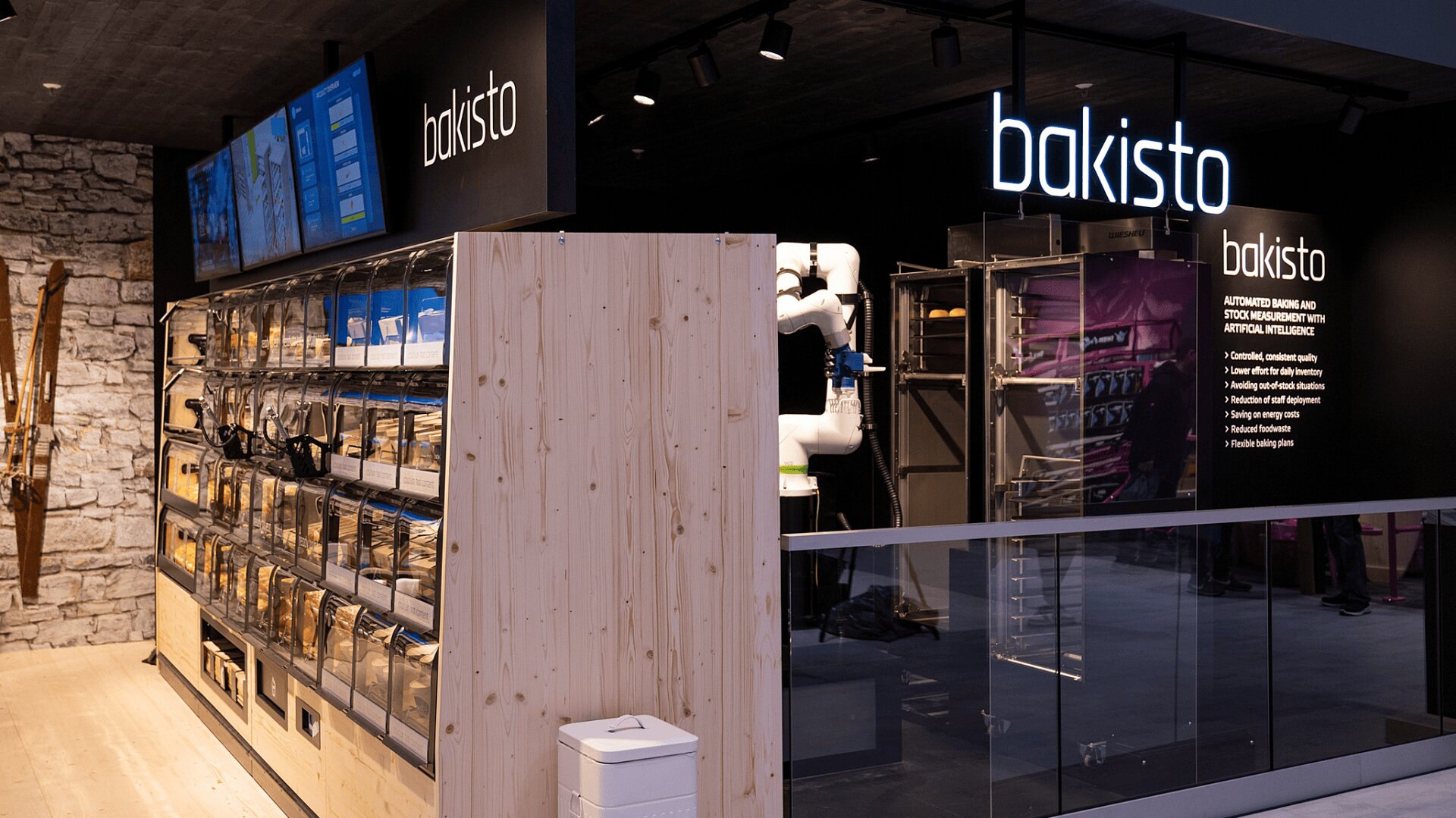 Eine moderne Selbstbedienungsbäckerei mit übersichtlichen Brotbehältern, einem Touchscreen und großen Bakisto-Schildern darüber. Industrielle Ausrüstung und Roboterarme sind hinter Glas im Hintergrund zu sehen.