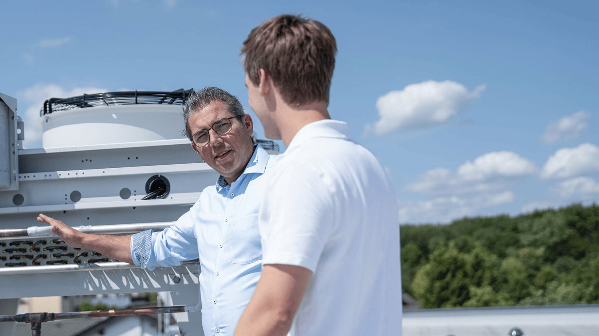Zwei Männer stehen auf einem Dach vor einer Industrieanlage und führen ein Gespräch. Der eine ist der Kamera zugewandt und gestikuliert mit seiner Hand, während der andere dem Betrachter den Rücken zuwendet. Im Hintergrund sind Bäume und ein blauer Himmel zu sehen.