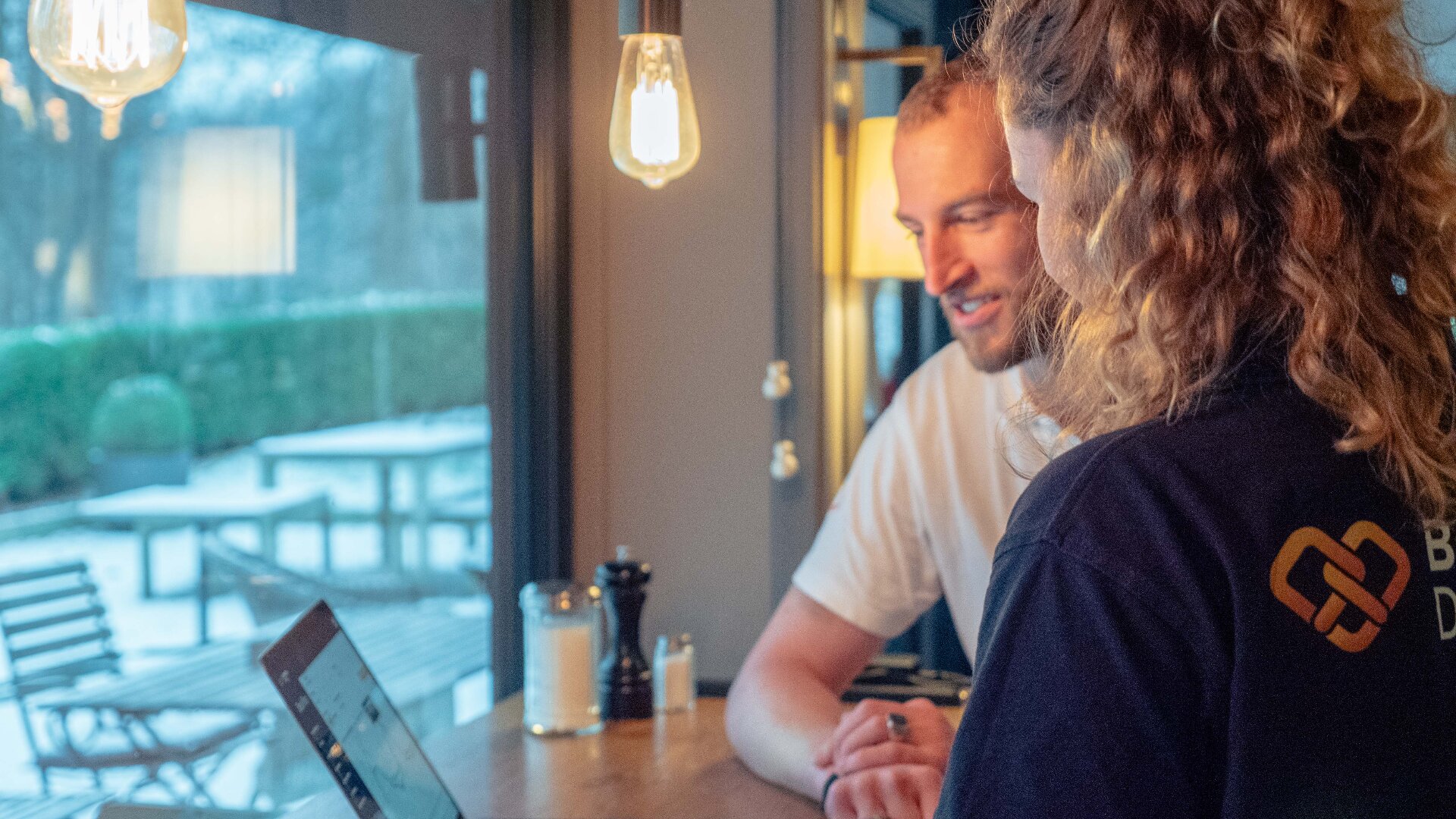 Zwei Personen sitzen an einem Holztisch am Fenster, schauen auf einen Laptop und lächeln. Durch das Fenster sind Sitzgelegenheiten im Freien und Grünflächen zu sehen. Die warme Innenbeleuchtung schafft eine gemütliche Atmosphäre.
