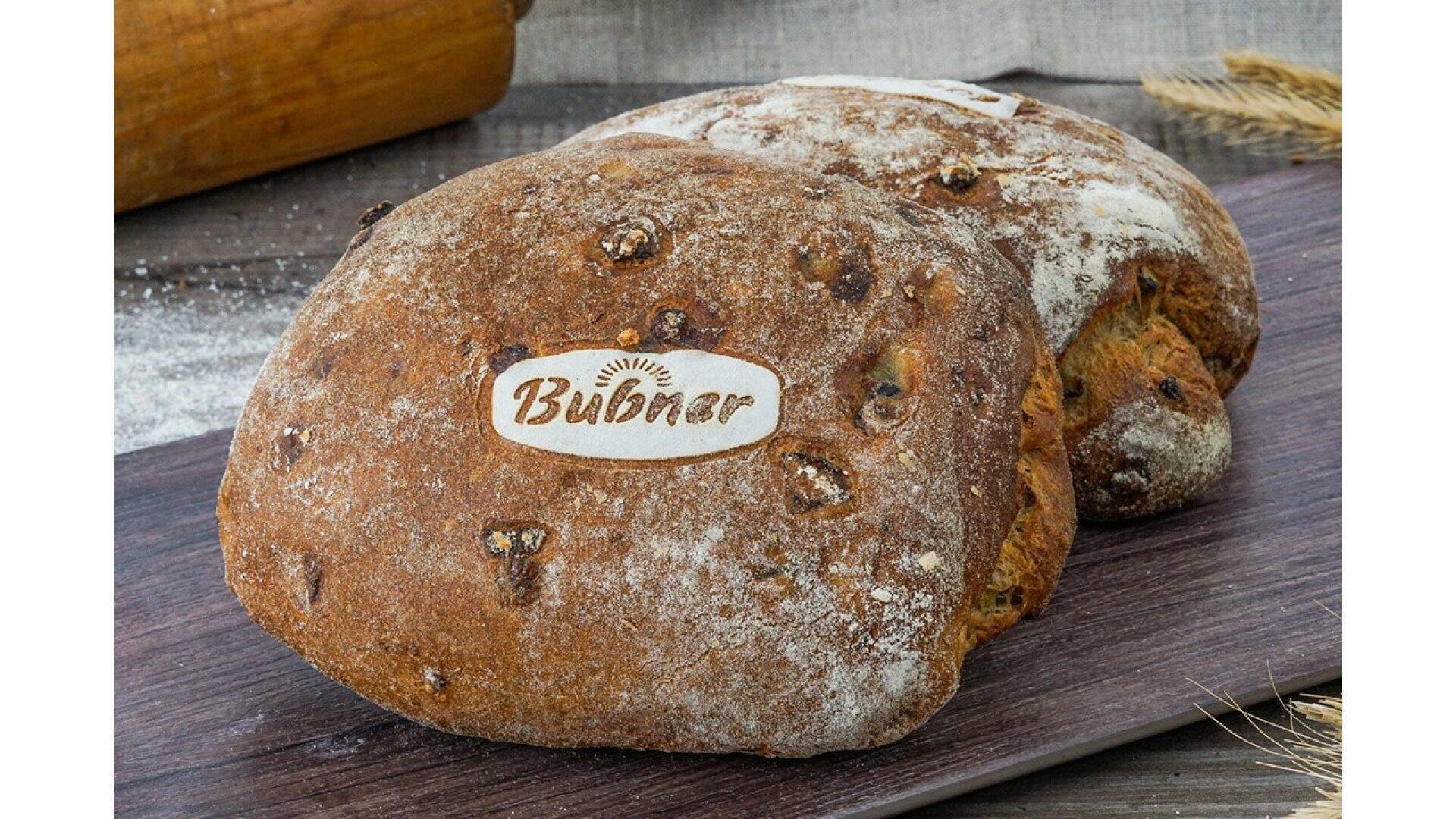 Two rustic loaves of bread with a dusting of flour and a “Bubnar” label on top, placed on a wooden board with some wheat stalks and a rolling pin in the background.