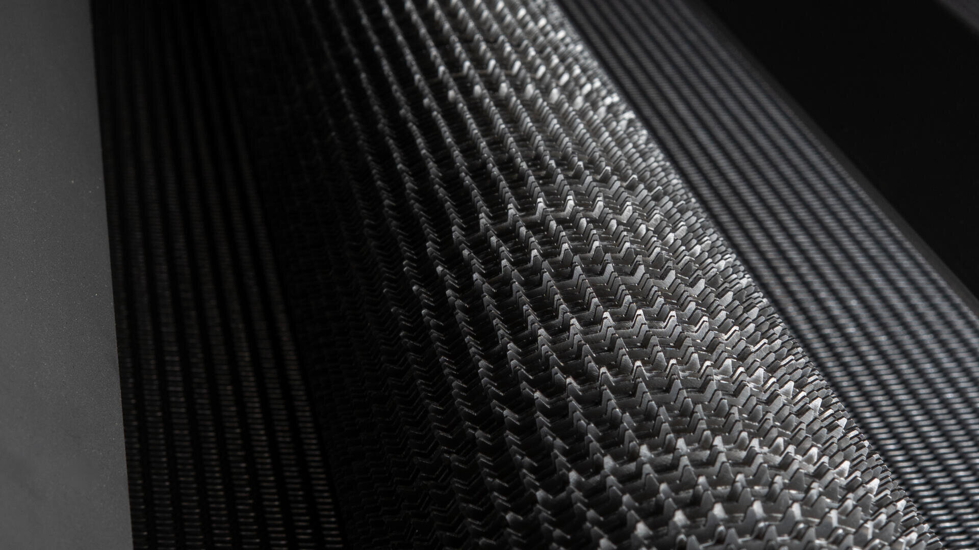 Nahaufnahme von verwobenem Kohlefasermaterial mit einem strukturierten Zickzackmuster. Die Beleuchtung hebt die komplizierte, reflektierende Oberfläche und die feine, detaillierte Struktur der Fasern hervor.