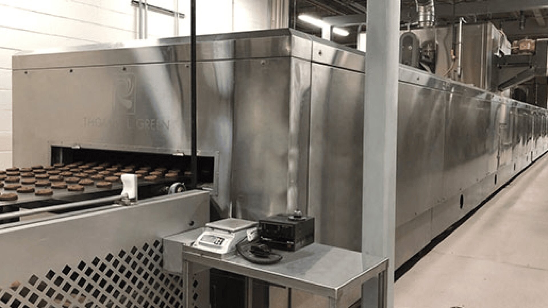 Large industrial baking oven with cookies on a conveyor belt entering the machine; digital scales and control equipment sit beside the oven in a clean, modern food production facility.