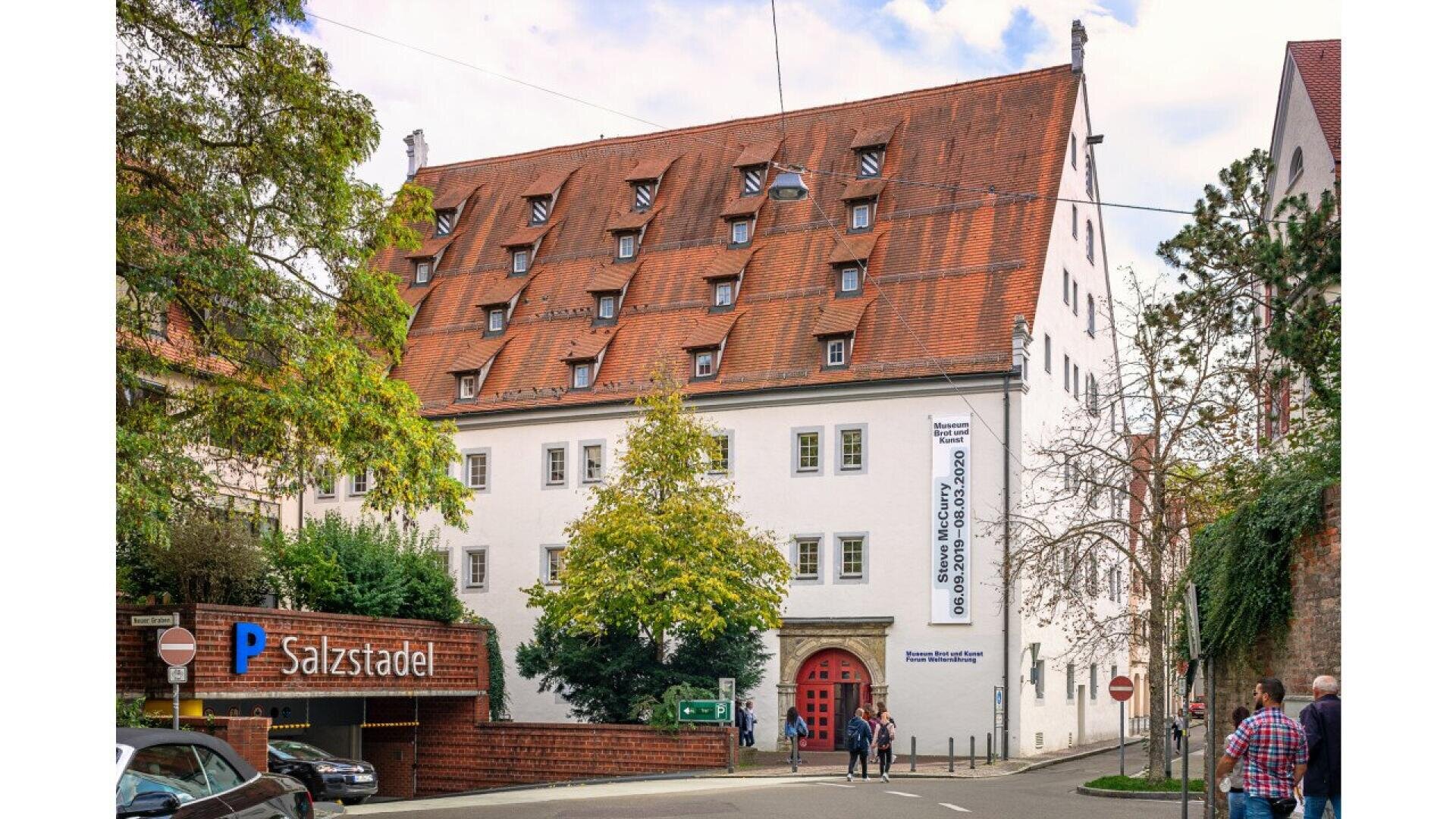 A historic white building with a steep red-tiled roof and many dormer windows stands behind trees. A banner hangs by the entrance. People walk nearby, and a Salzstadel parking sign is visible in the foreground.