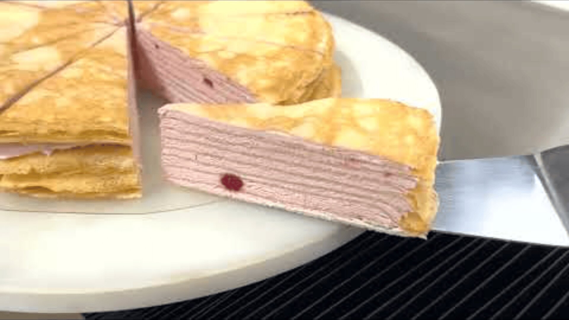 A slice of layered crepe cake with a light pink filling and a single red spot is lifted out of the whole cake on a white plate.