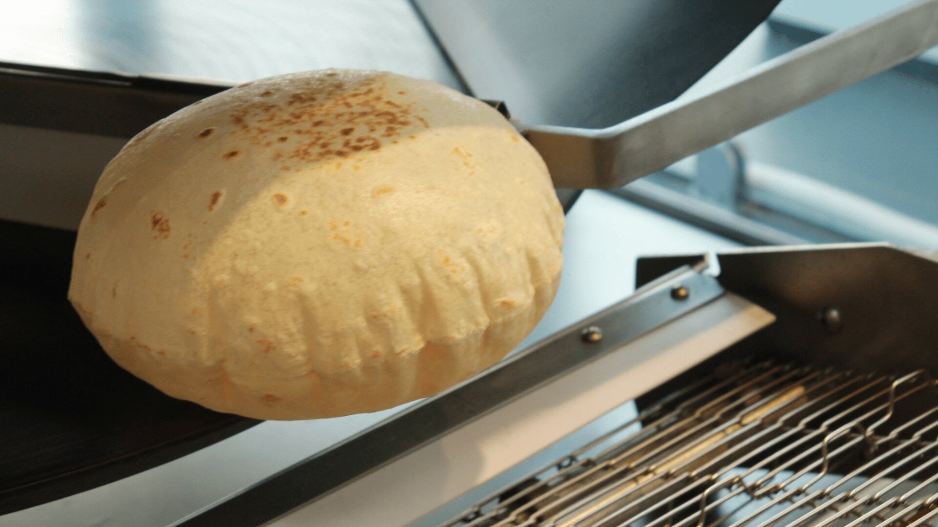A freshly baked, round flatbread is puffed up and resting on a metal baking machine or conveyor in a kitchen or bakery setting.