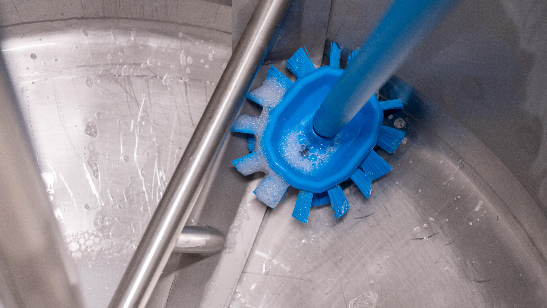 Eine Nahaufnahme einer blauen Reinigungsbürste, die das Innere eines großen Metallbehälters schrubbt, wobei etwas Wasser und Seifenlauge zu sehen sind.