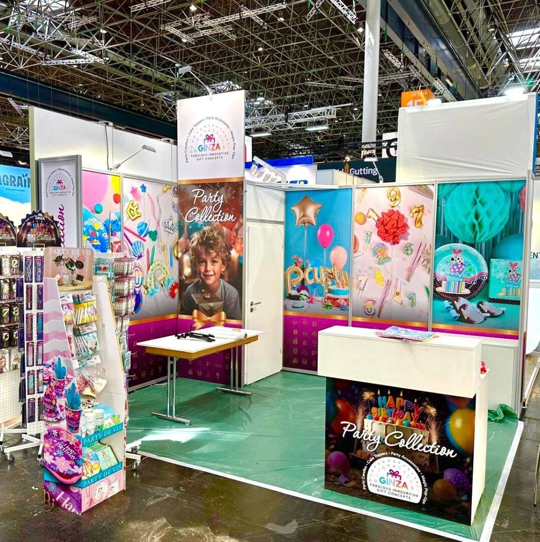 Ein farbenfroher Messestand präsentiert Partyzubehör wie Luftballons, Banner und Dekorationen. Bunte Poster, ein lächelndes Kind und geordnete Produkte sorgen für festliche Atmosphäre. Auf einem Tisch und in Regalen finden sich weitere Partyartikel.