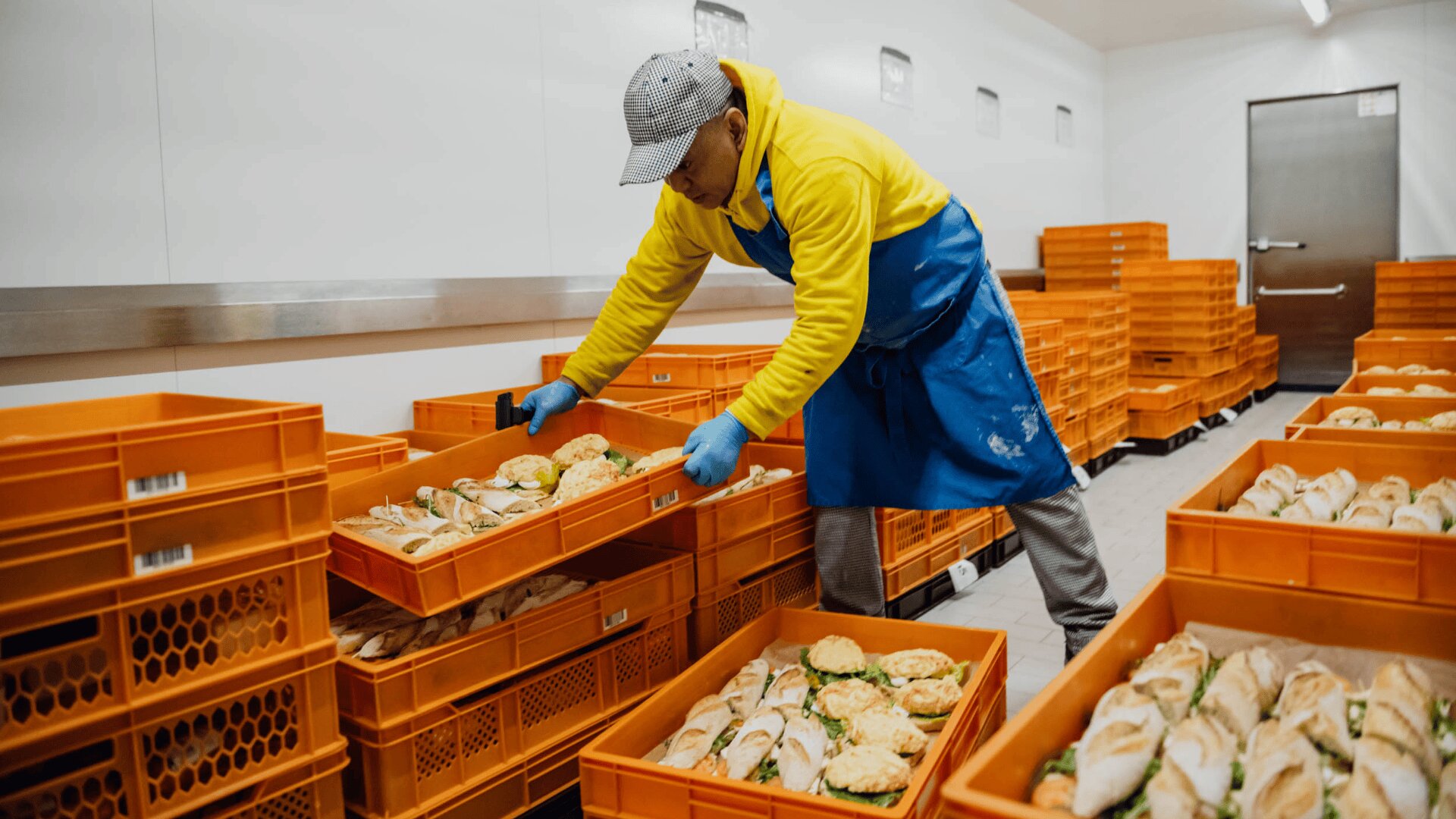 Ein Arbeiter mit gelbem Kapuzenpulli, blauer Schürze und Handschuhen stapelt orangefarbene Kisten mit Sandwiches oder Brötchen in einem Lebensmittellager oder Zubereitungsbereich.