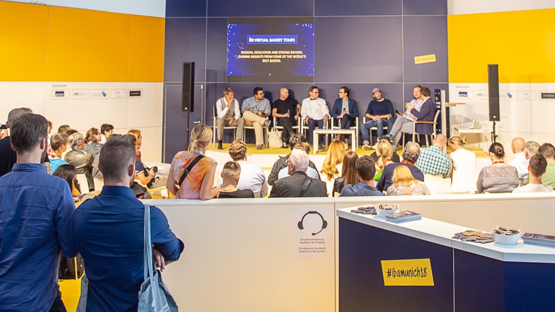Eine Gruppe von sieben Rednern sitzt auf der Bühne vor dem Publikum einer Konferenz und diskutiert über ein Thema, das auf einem Bildschirm hinter ihnen angezeigt wird. Das Publikum hört aufmerksam zu. Die Umgebung ist modern mit leuchtend gelben und blauen Wänden.