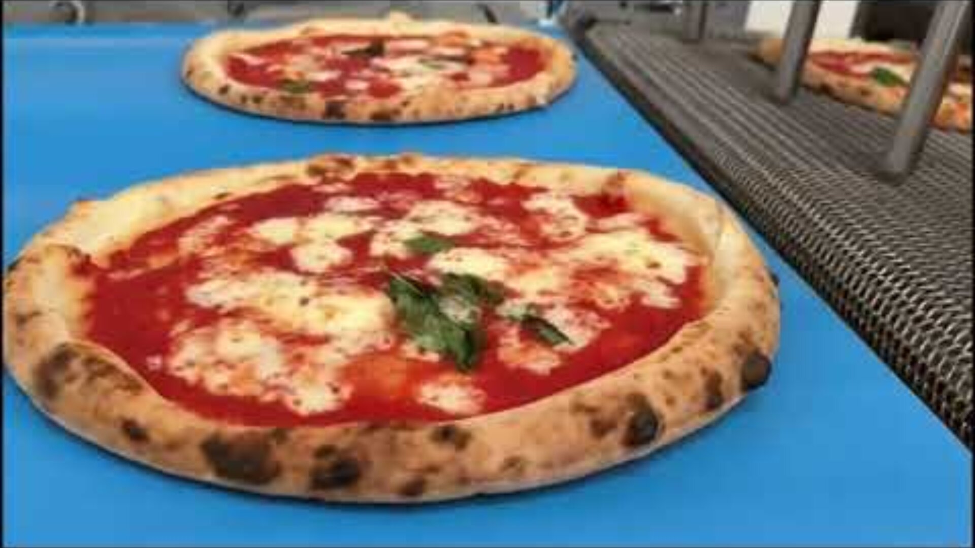 A close-up of a freshly baked Margherita pizza with melted cheese, tomato sauce, and basil leaves on a blue conveyor belt, with another pizza in the background.