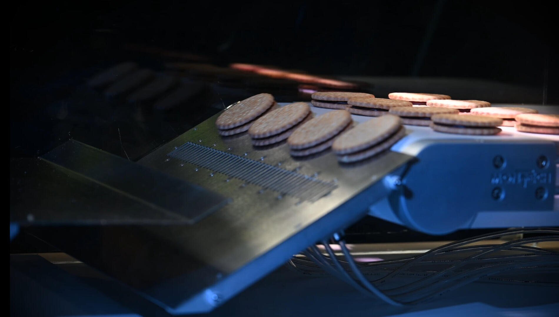 Nahaufnahme eines Förderbands, das bei schwacher Beleuchtung Reihen von Sandwich-Keksen durch eine Fertigungsmaschine transportiert.