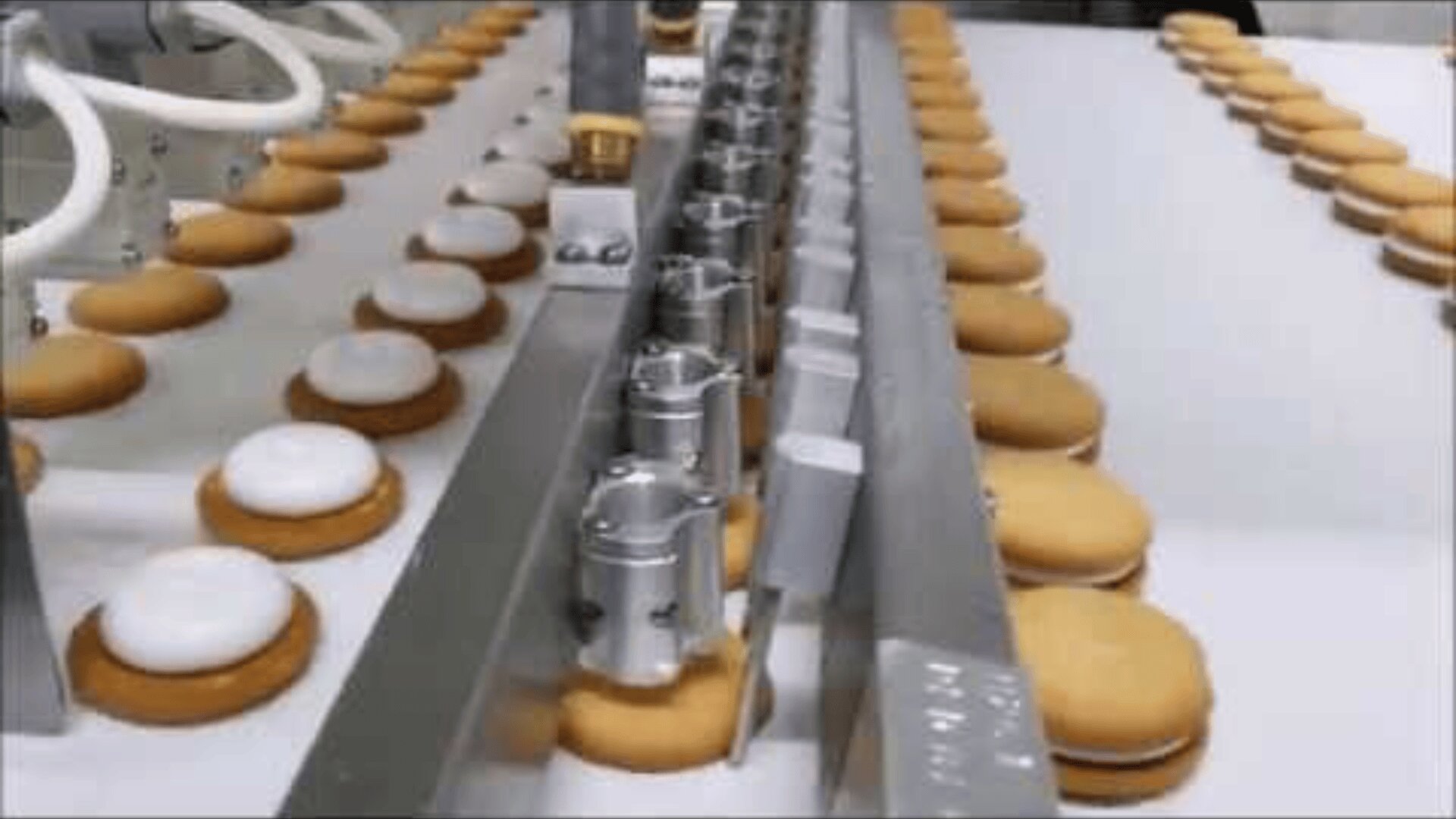 In a factory, cookies run along a conveyor belt. Some cookies are covered in white icing, others are unprinted, and a machine appears to be assembling or decorating them.