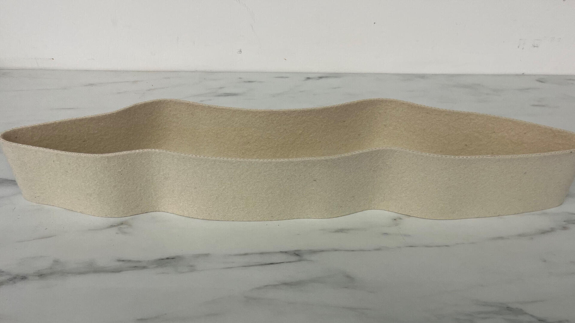 Ein beiger, gewellter, flacher Filzkorb steht auf einer weißen Marmorfläche vor einem schlichten weißen Hintergrund.