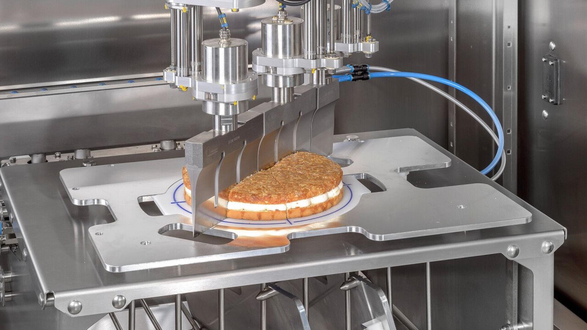 A machine uses multiple blades to evenly slice a round sandwich or pastry on a metal tray in a stainless steel industrial food processing environment.