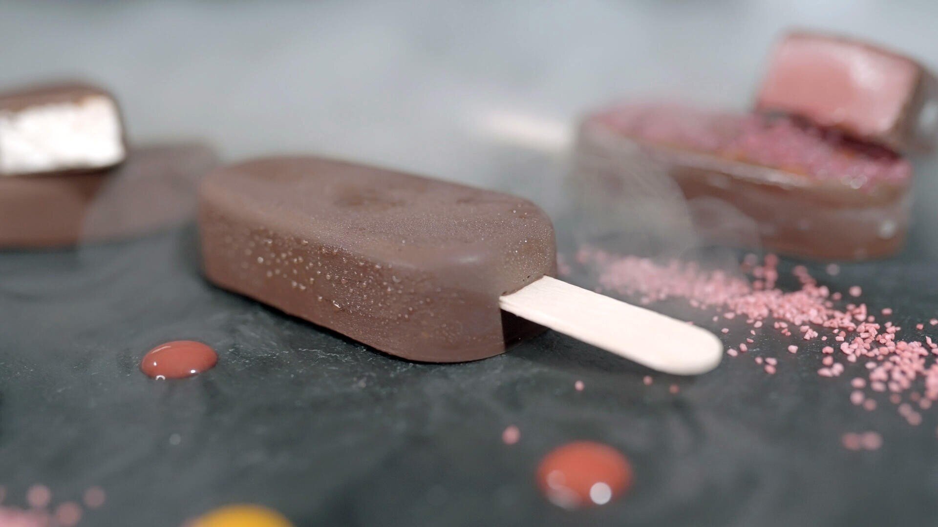 A chocolate-coated ice cream bar on a stick rests on a dark surface, surrounded by red and yellow syrup drops and pink sprinkles, with a blurred dessert in the background.