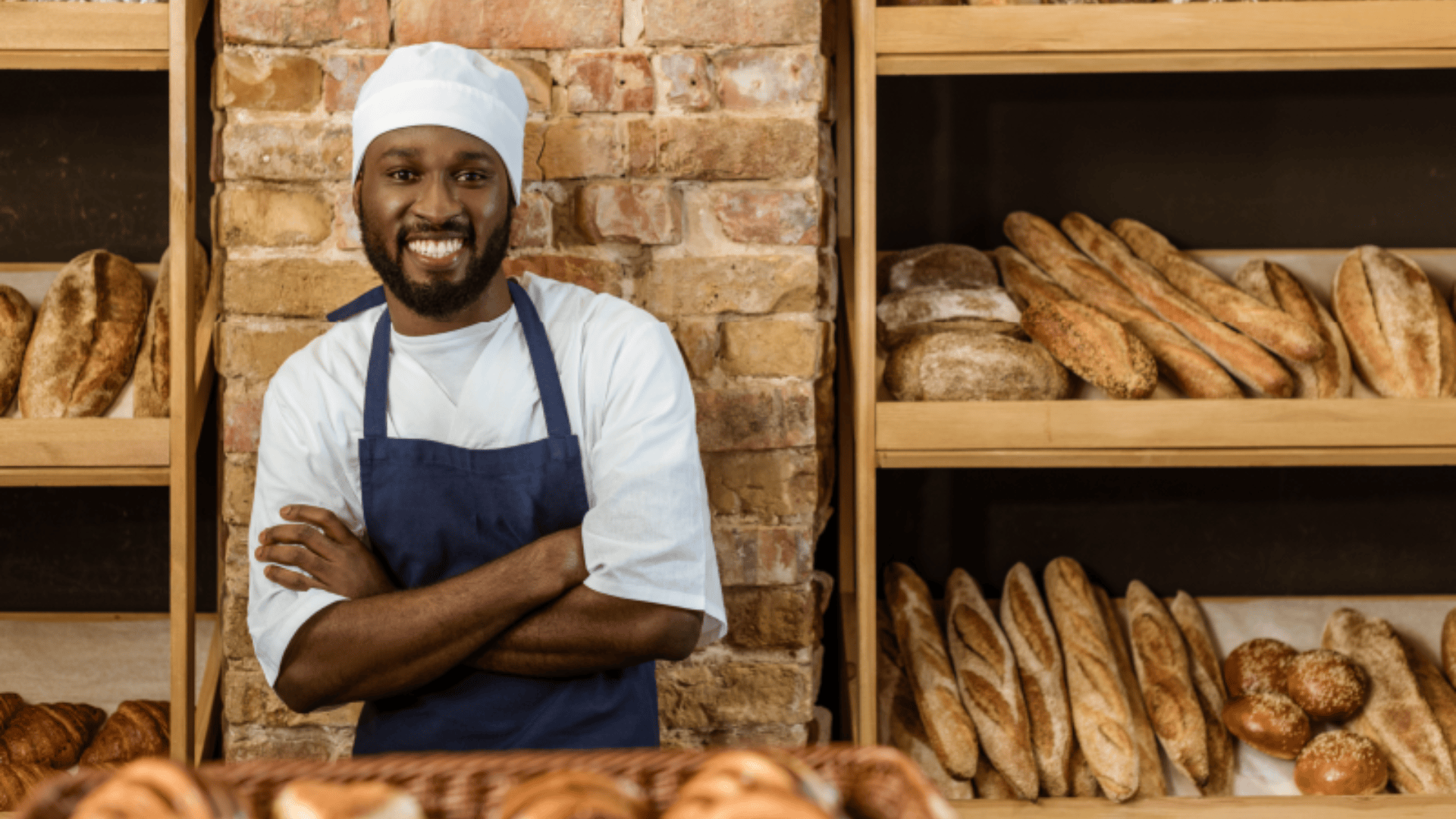 A smiling baker wearing a white hat and blue apron stands with arms crossed in front of shelves filled with baguettes and other bread in a rustic bakery.