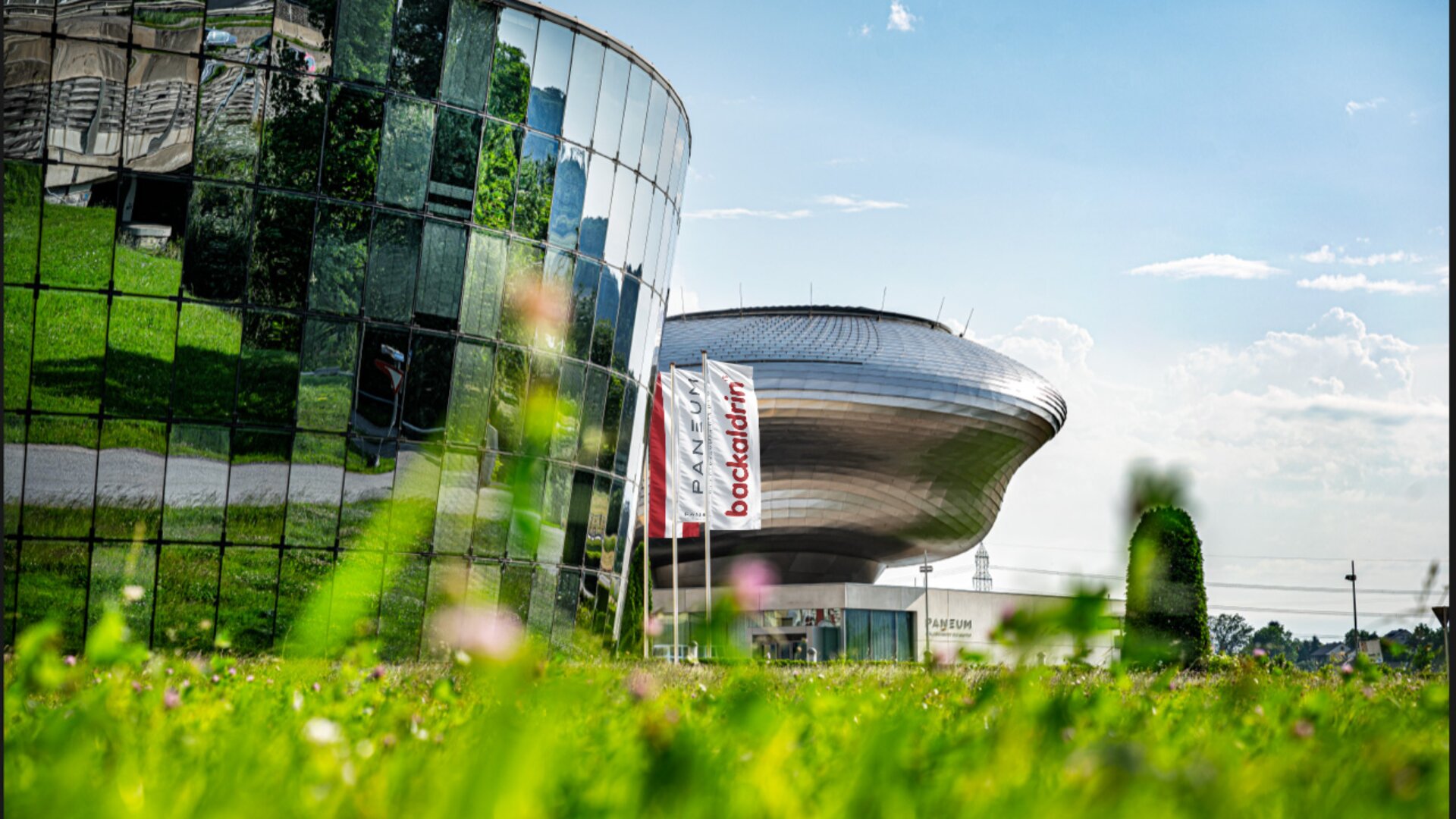 Ein modernes, geschwungenes Glasgebäude, in dem sich Grün spiegelt, steht neben einer futuristischen, silbernen Struktur. Dazwischen stehen Fahnen mit rotem Text, und das Bild ist von einem grasbewachsenen, mit Blumen übersäten Rasen unter blauem Himmel aufgenommen.