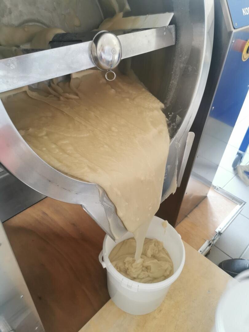 Ein dicker, cremiger Teig fließt aus einem großen industriellen Mixer in einen weißen Plastikeimer, der unten in einer Küche oder Lebensmittelproduktion steht.