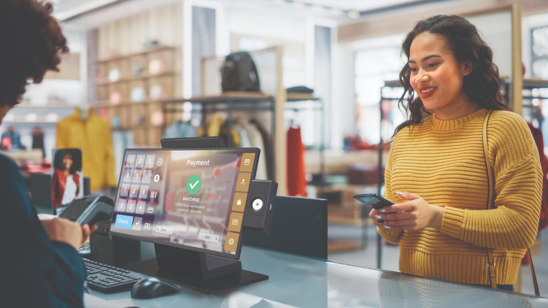 Eine Frau in einem gelben Pullover lächelt, während sie an einer Ladentheke eine mobile Zahlung tätigt. Ihr Gesicht steht einer Kassiererin gegenüber. Zwischen ihnen ist auf der Kasse ein Bildschirm mit einer digitalen Zahlungsbestätigung zu sehen.