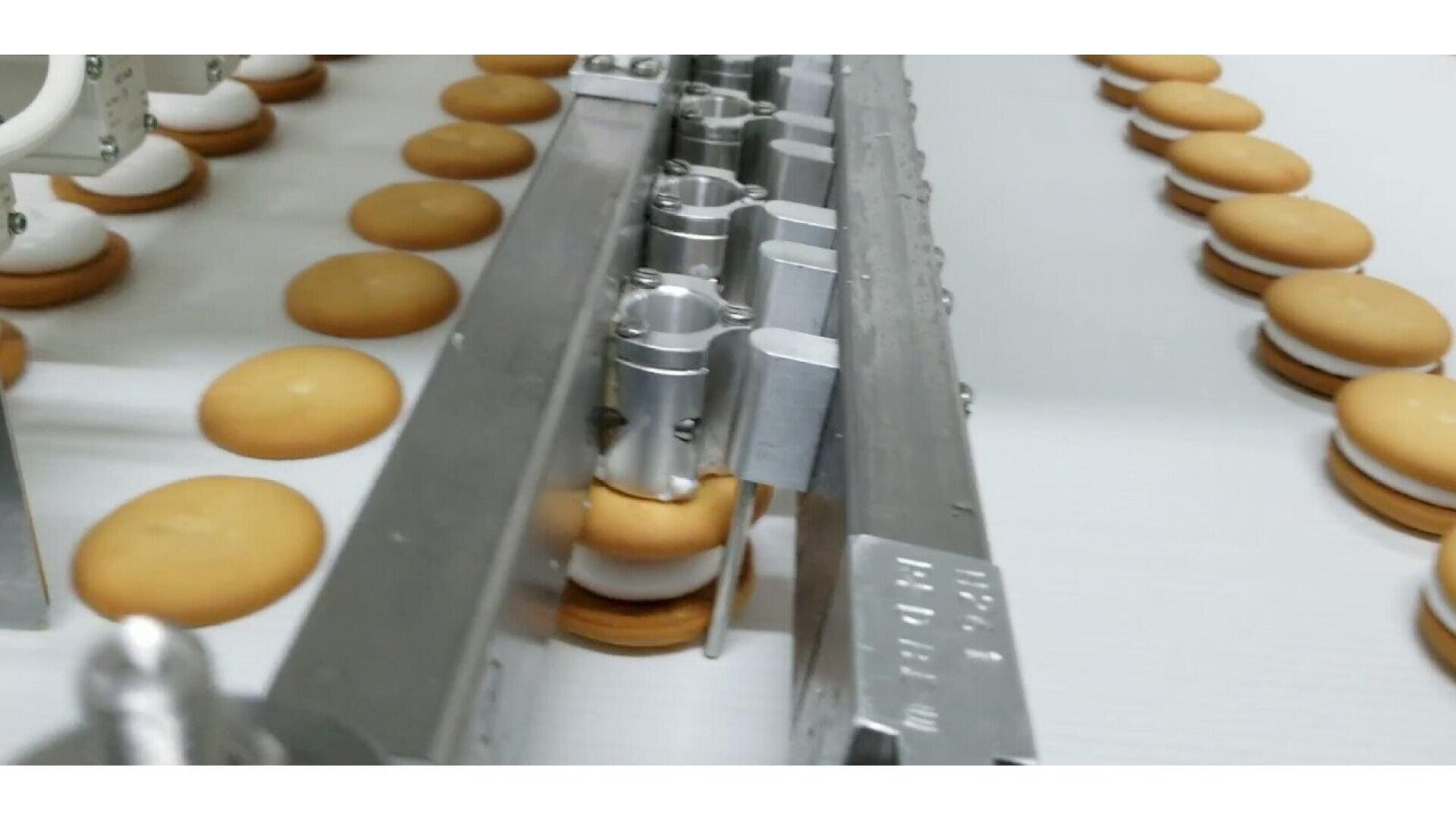Nahaufnahme einer Lebensmittelproduktionslinie, in der runde Sandwich-Kekse mit weißer Cremefüllung zusammengesetzt und über ein Förderband transportiert werden.