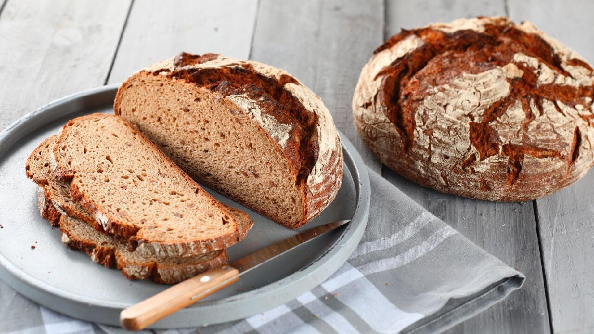 A round loaf of rustic brown bread sits on a wooden table next to a plate with several slices of the same bread and a knife, with a gray striped napkin underneath.