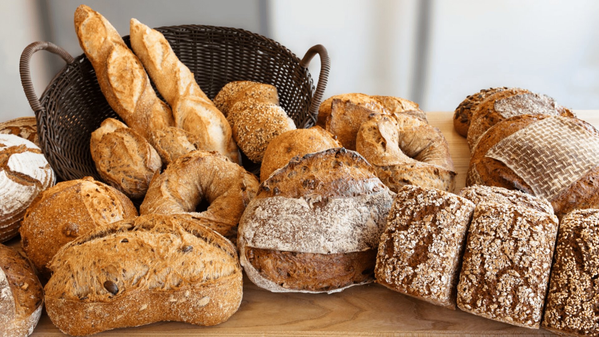 Verschiedene handwerklich hergestellte Brotsorten, darunter Baguettes, runde Brote, Körnerbrötchen und rustikales Brot, werden auf einer Holzfläche mit einem dunklen geflochtenen Korb im Hintergrund präsentiert.