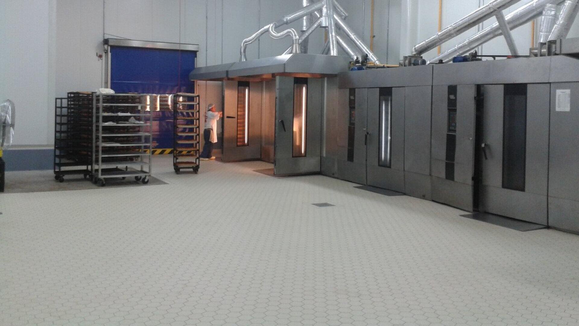 Eine gewerbliche Bäckerei mit großen Industrieöfen, Metallgestellen auf Rädern und einem Arbeiter im weißen Kittel, der teilweise durch eine offene Ofentür sichtbar ist. Der Boden ist gefliest, und an der Decke sind Lüftungsschlitze angebracht.