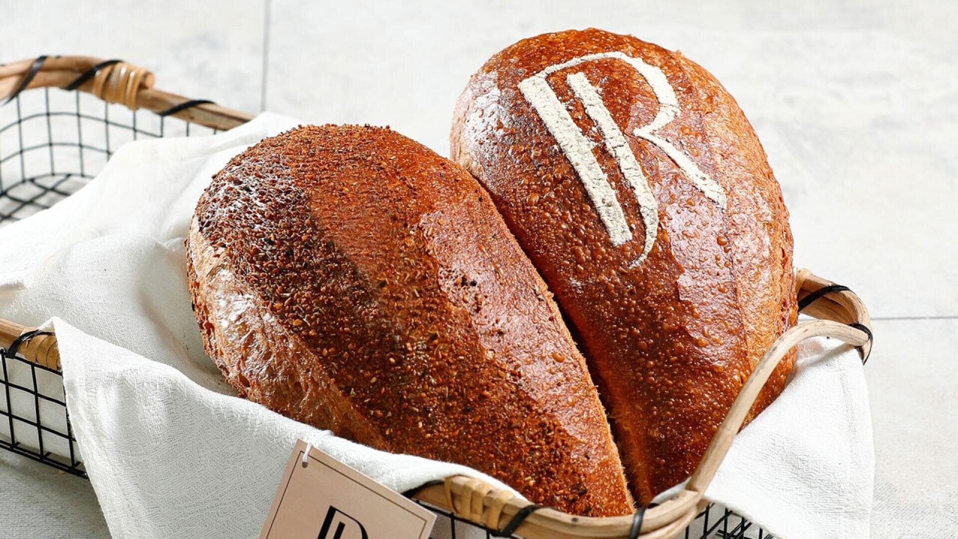 Two loaves of brown bread in a basket lined with a white cloth, one loaf topped with seeds and the other with a white JR logo stenciled on the crust.