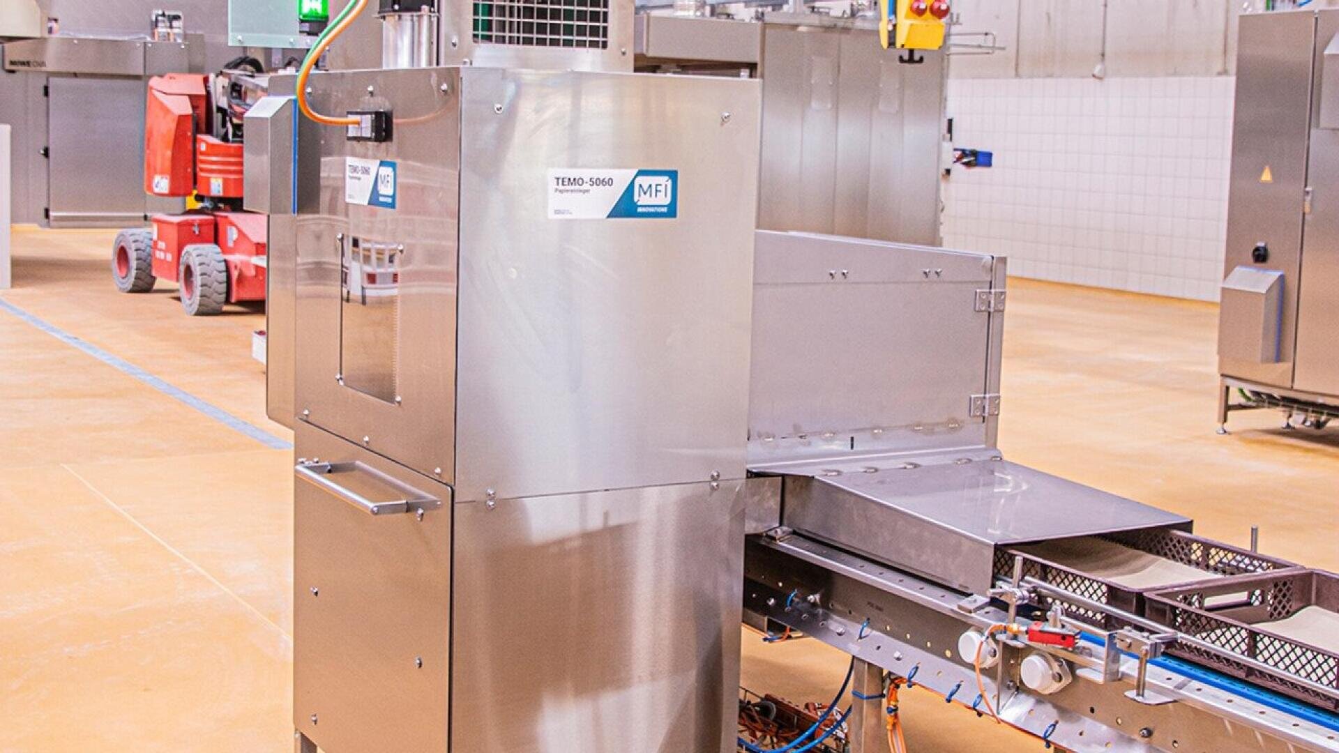 Eine Industriemaschine aus rostfreiem Stahl mit einem Förderband in einer sauberen, geräumigen Fabrikumgebung. Die Maschine hat ein Bedienfeld und ist mit TEMPO 5000 beschriftet. Im Hintergrund sind weitere Geräte zu sehen.