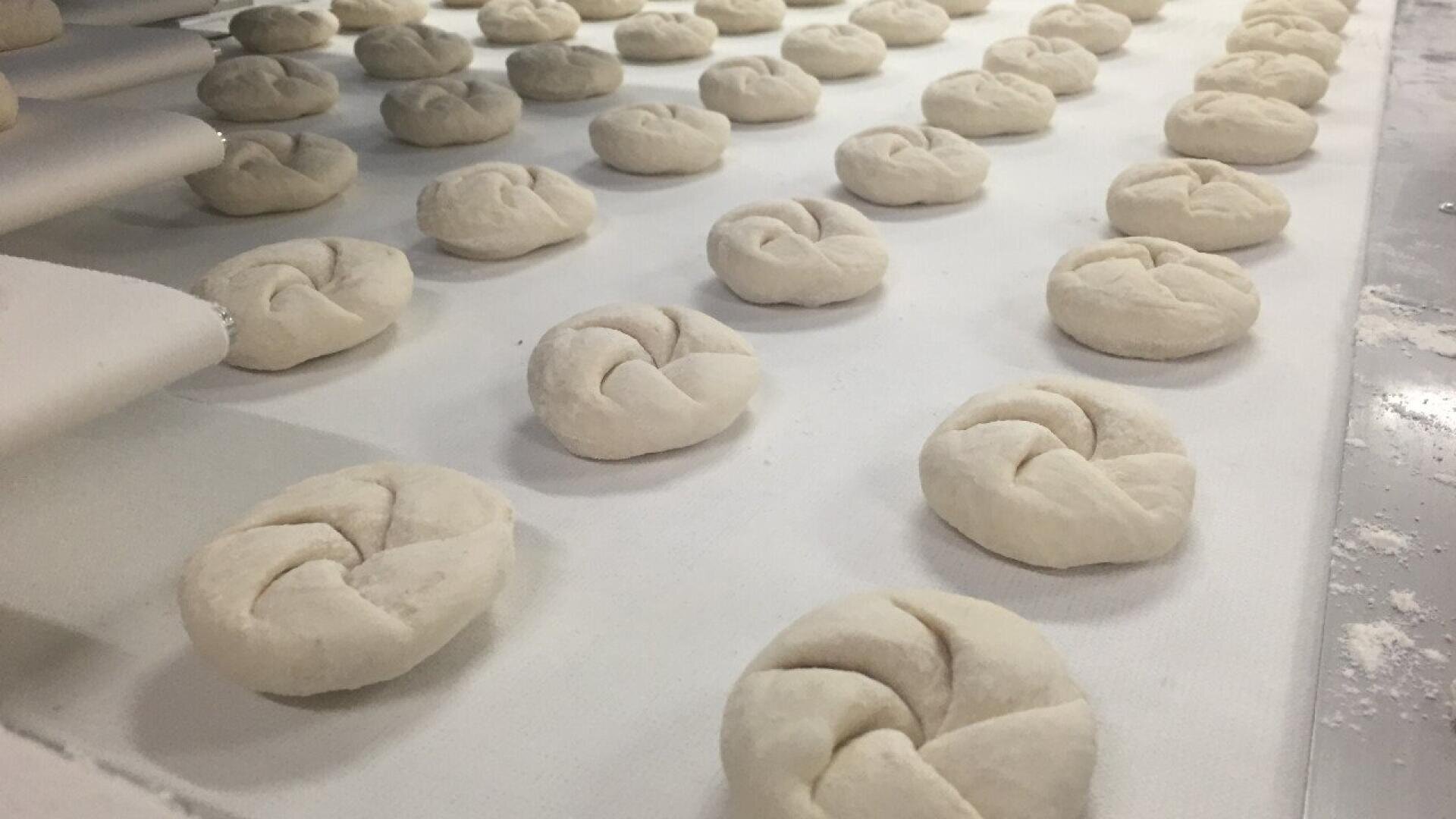 Die ungebackenen, verknoteten Teigrollen werden in Reihen auf einer bemehlten Fläche angeordnet und sind bereit zum Backen. Die Teigstücke liegen gleichmäßig verteilt und sind leicht mit Mehl bestäubt.