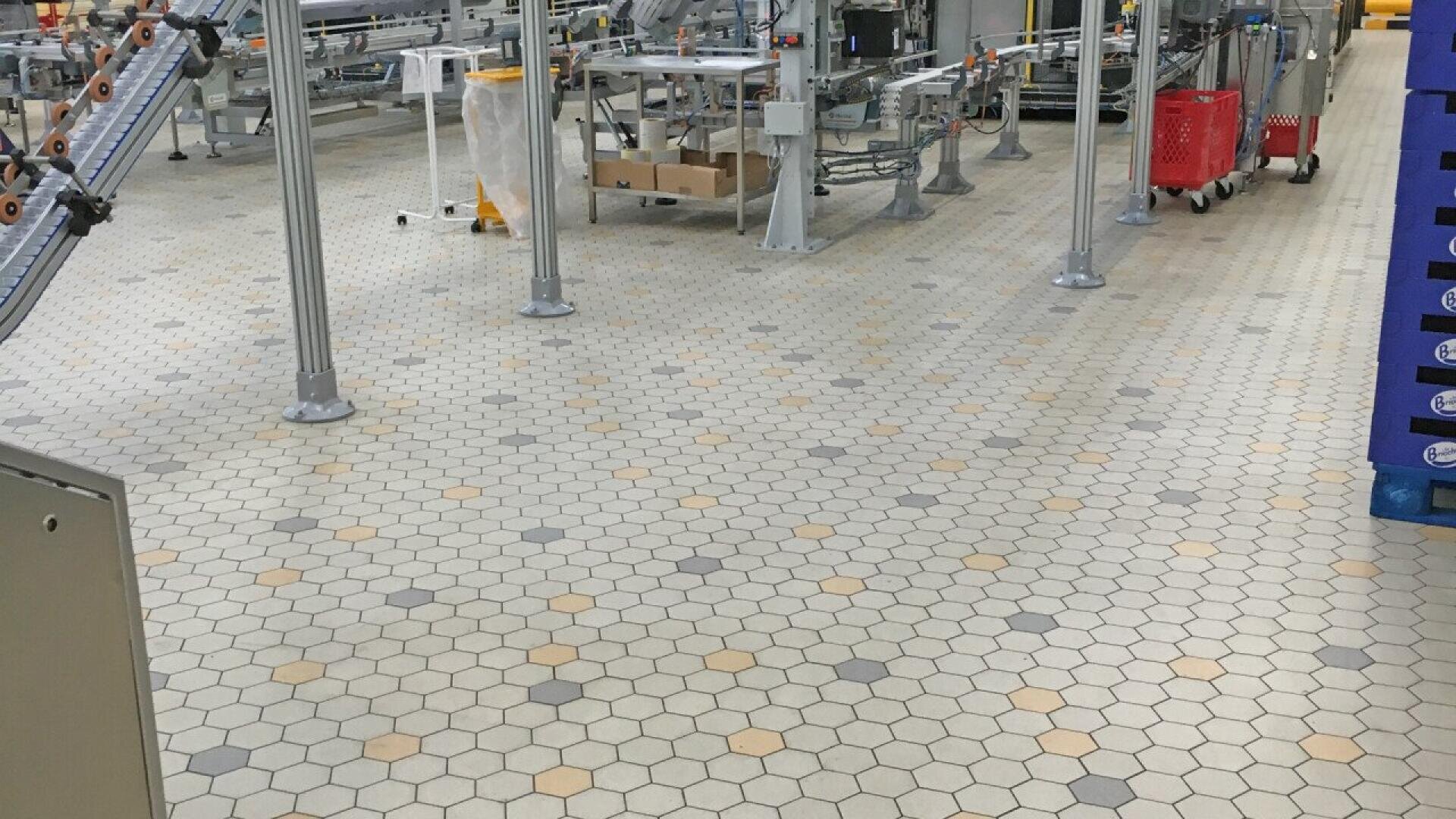 A factory or industrial workspace with hexagonal tile flooring in beige, gray, and yellow tones; visible are metal posts, conveyor belts, machinery, and red and blue containers in the background.
