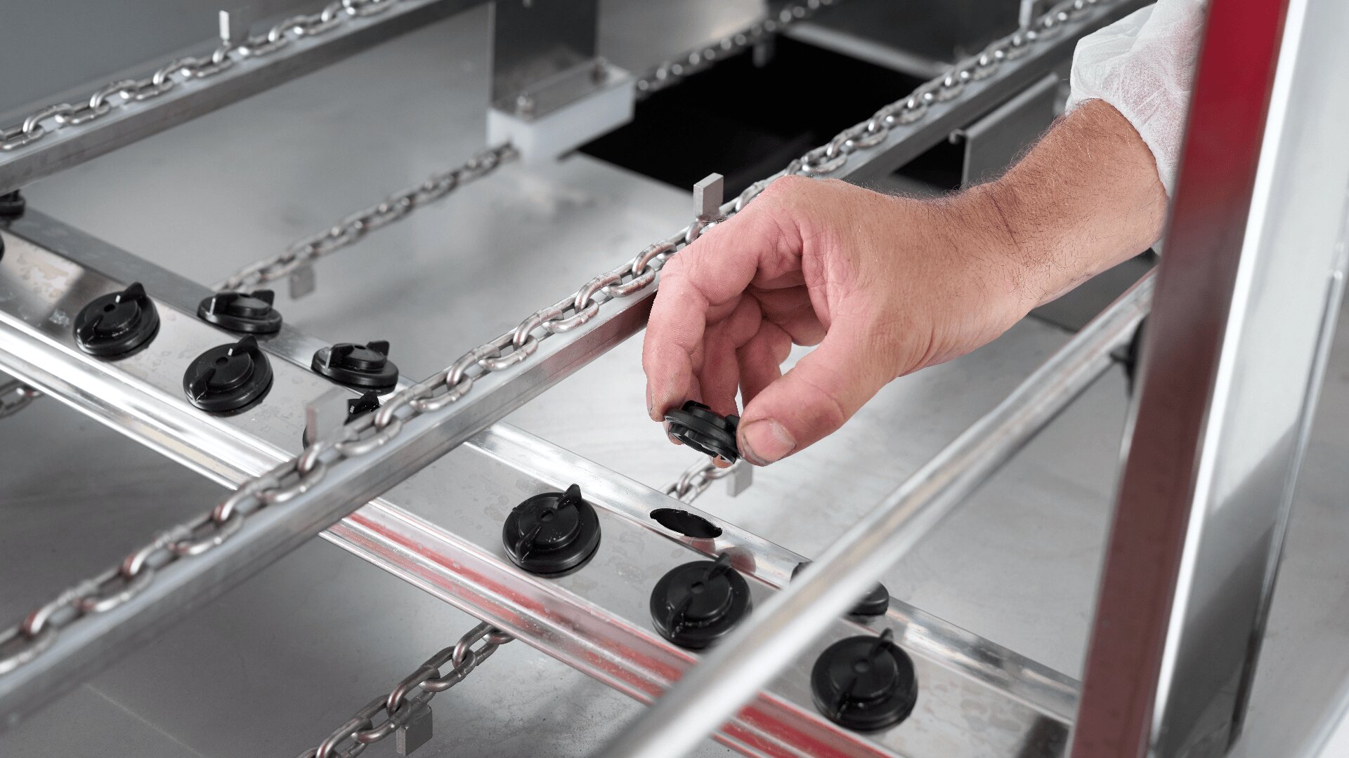 Nahaufnahme der Hand einer Person, die schwarze Kappen an einer Metallproduktions- oder Montagelinie einstellt oder prüft, wobei Ketten und Metallschienen in einer industriellen Umgebung sichtbar sind.