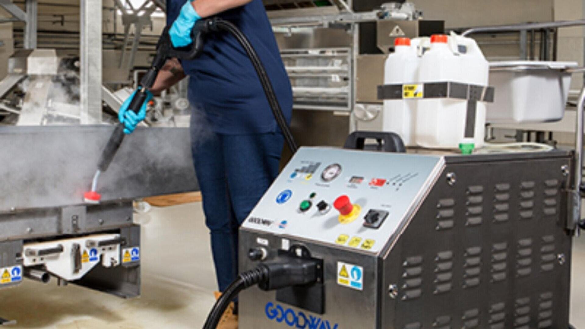 Eine Person mit blauen Handschuhen benutzt eine industrielle Dampfreinigungsmaschine, um Fabrikanlagen zu desinfizieren; die Maschine ist mit Kontrollknöpfen und Warnschildern versehen, und in der Nähe stehen Plastikbehälter mit Chemikalien.