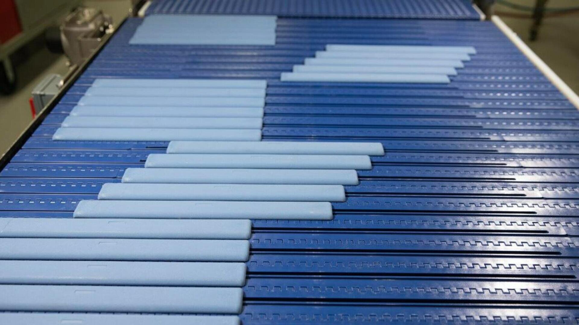Blaues Förderband mit gleichmäßig verteilten, rechteckigen, hellblauen Stäben, die in diagonalen Reihen angeordnet sind und sich entlang einer industriellen Verarbeitungslinie bewegen.