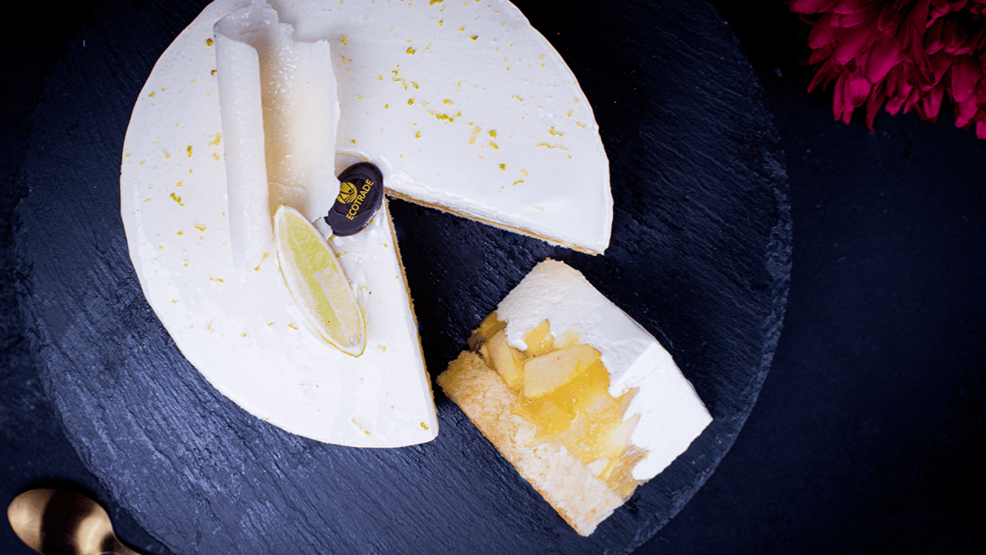 Ein runder, weißer, mit Limetten und weißer Schokolade garnierter Kuchen liegt auf einer dunklen Schiefertafel. Eine Scheibe ist herausgeschnitten und gibt den Blick frei auf Schichten von Sahne und Fruchtfüllung im Inneren. Ein goldener Löffel und eine rote Blume liegen daneben.