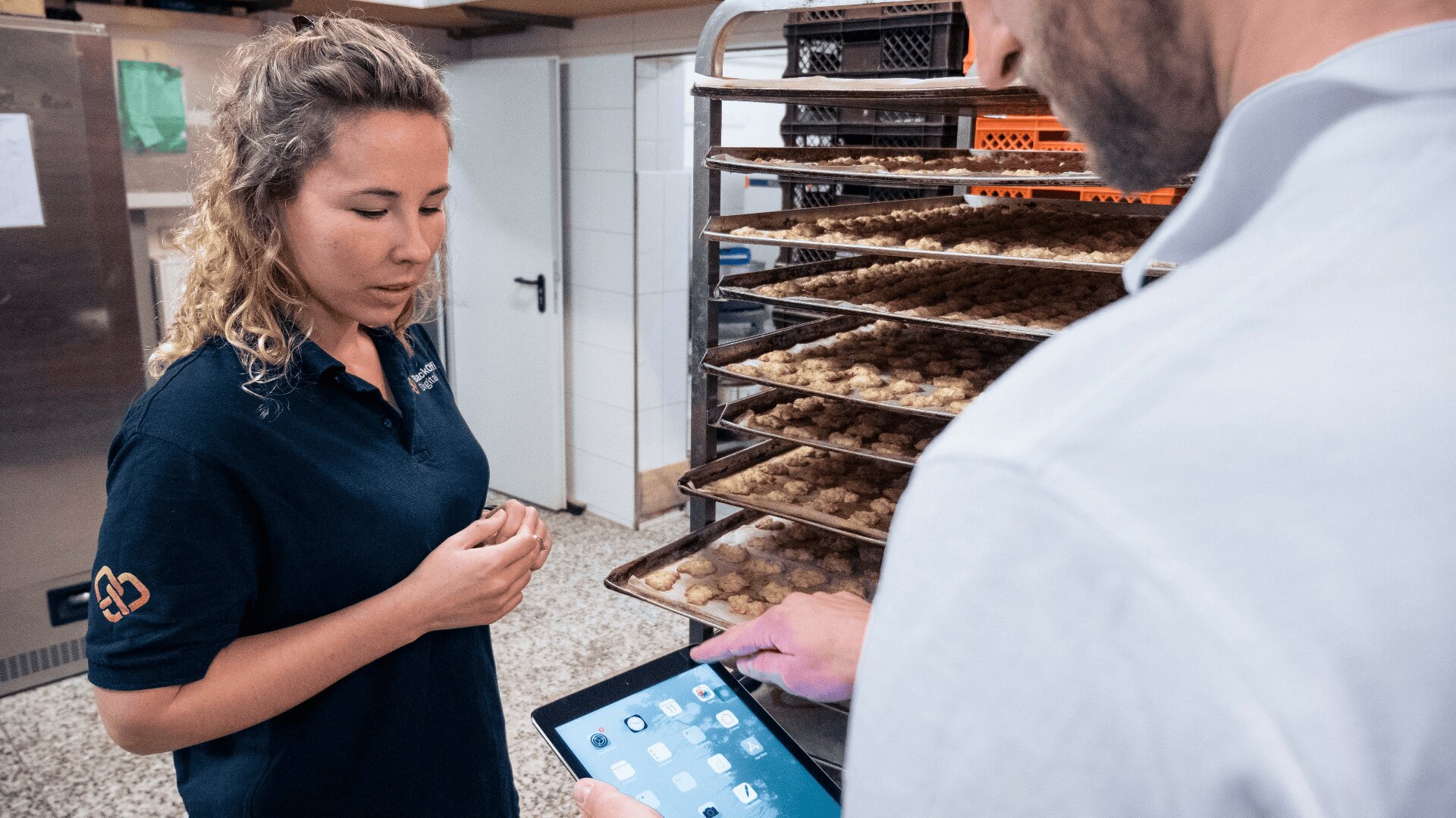 Eine Frau steht neben Tabletts mit Keksen auf einem Regal, während ein Mann ein Tablet benutzt und etwas in einer Großküche bespricht oder überprüft.