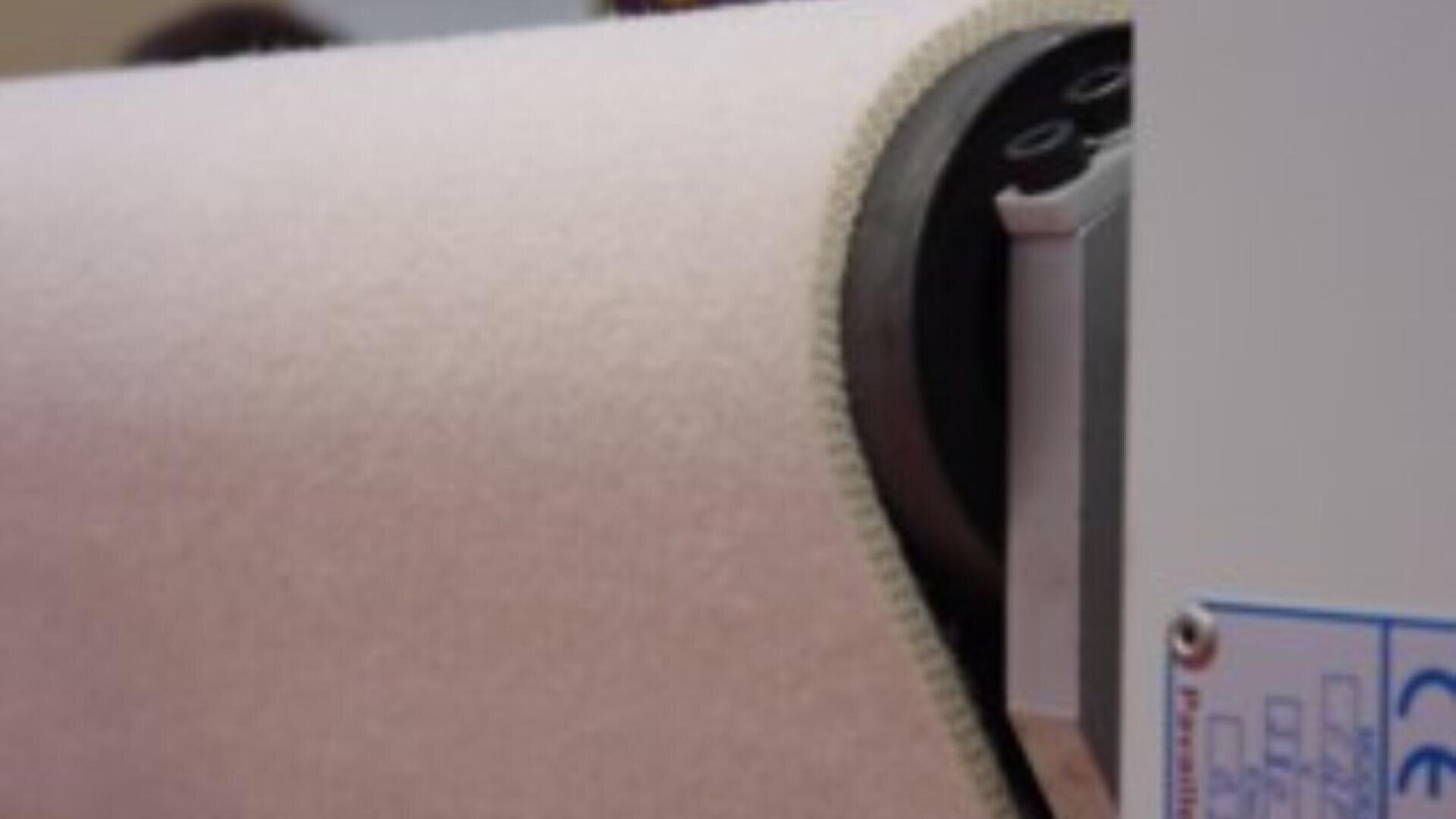 A close-up of a large roll of white fabric being processed by an industrial machine, with part of the machine and control panel visible on the right side.