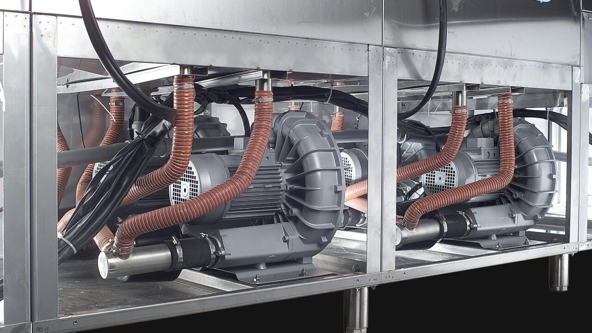 Zwei große elektrische Industriegebläse mit orangefarbenen Spiralschläuchen sind nebeneinander in einem Metallgehäuse installiert, wobei verschiedene Kabel und Rohre mit den Geräten verbunden sind.