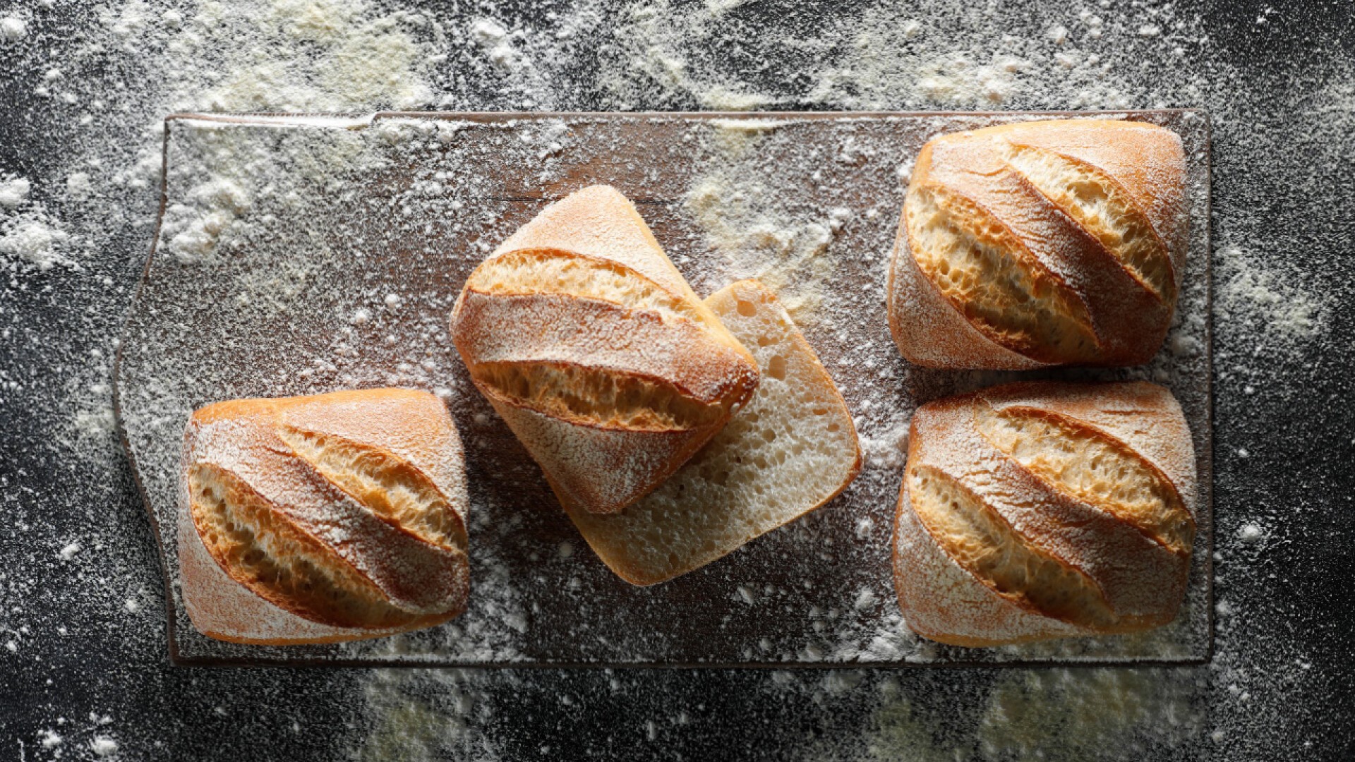 Four square pieces of rustic bread with golden crusts and flour dusting are arranged on a glass cutting board, with flour scattered on the dark surface around them.