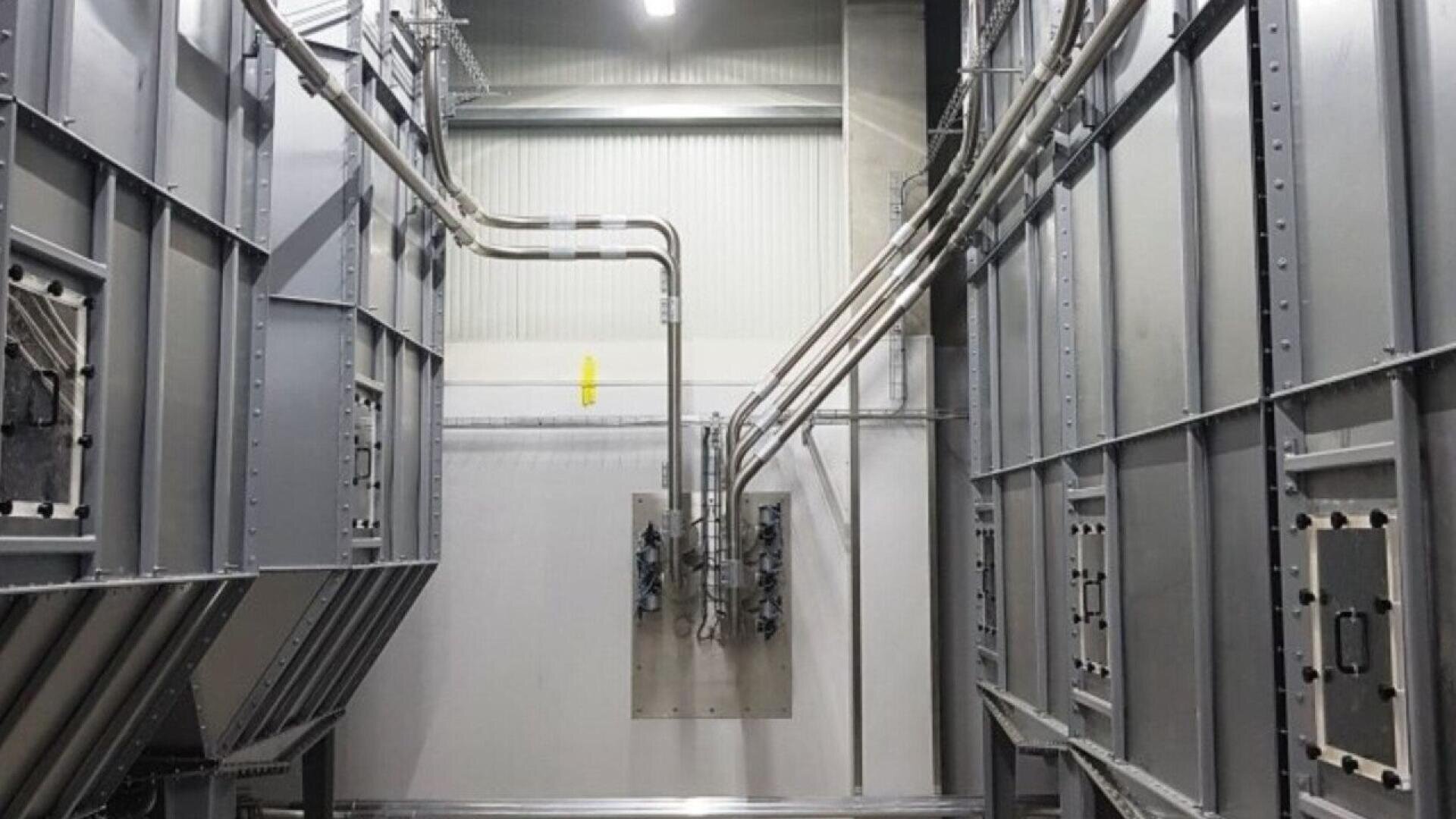 Interior view of an industrial facility with large metal structures on both sides, exposed pipes and cables running overhead and down to a wall-mounted panel, and bright overhead lighting.