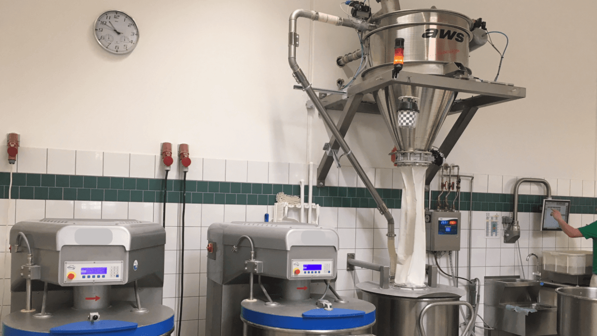 A modern industrial kitchen with large stainless steel machines, a digital control panel, a clock on the wall, and a complex ingredient dispensing system mounted above mixing bowls.