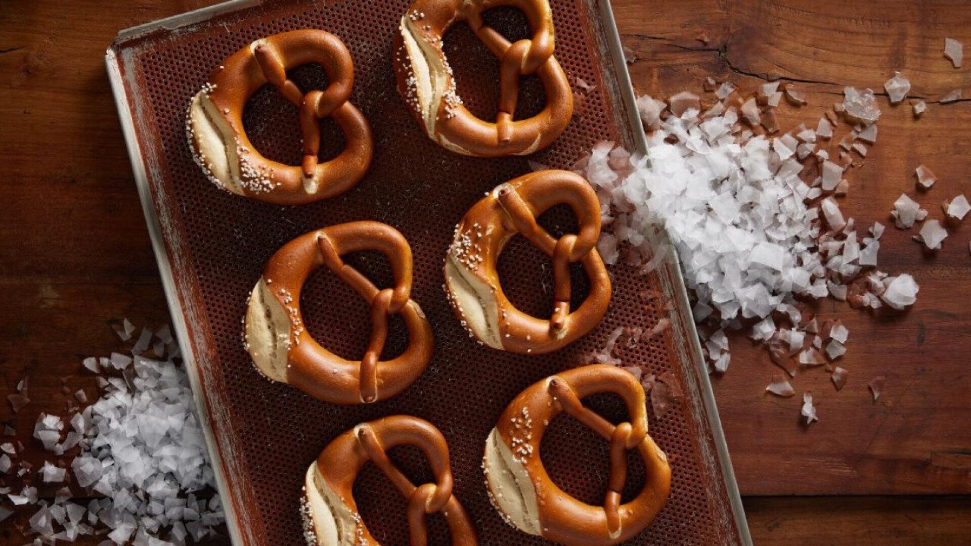 A baking sheet holds six golden-brown soft pretzels sprinkled with coarse salt, surrounded by scattered salt flakes on a wooden surface.