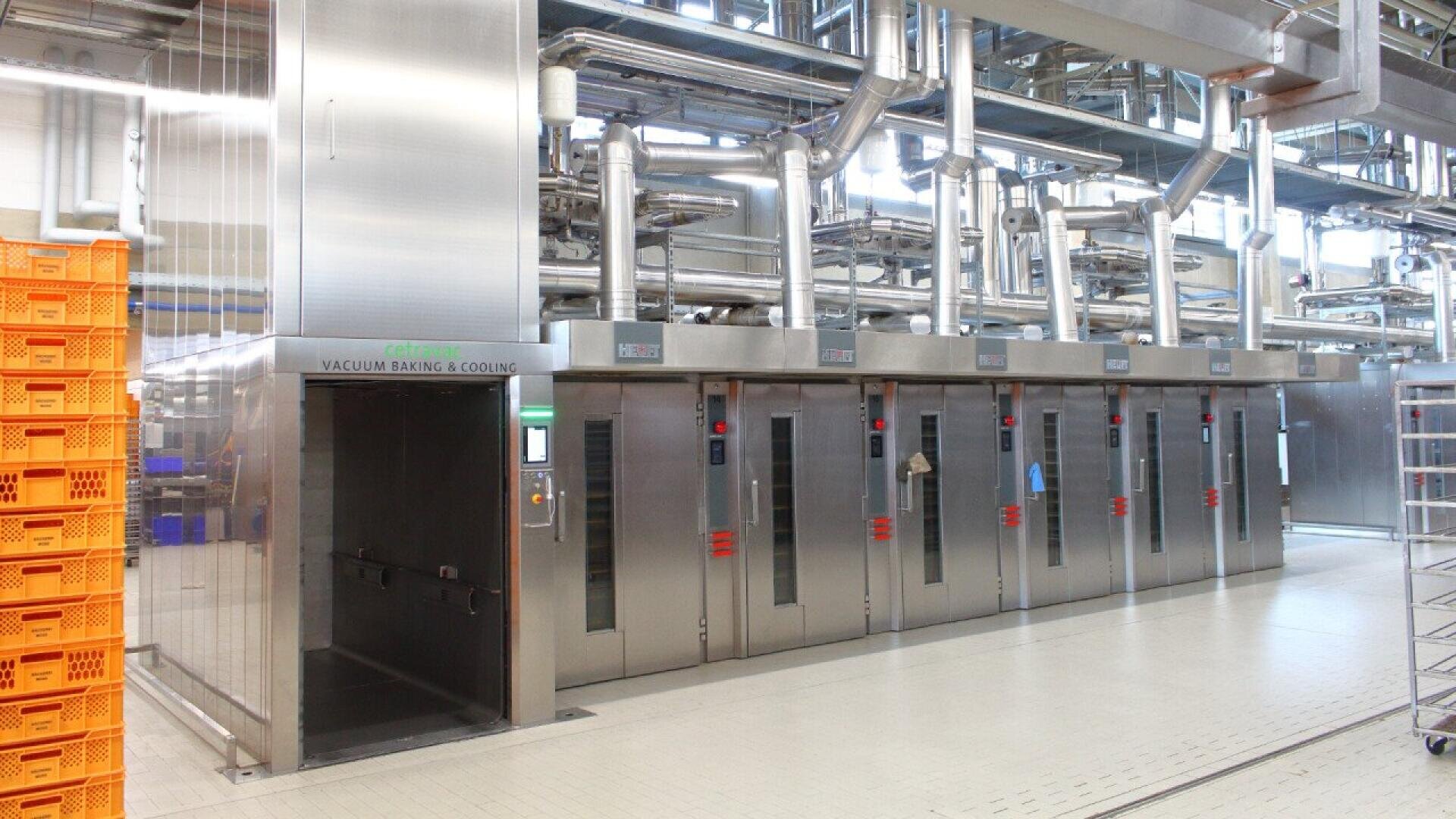 Eine moderne Industriebäckerei mit Öfen aus rostfreiem Stahl, Schalttafeln und umfangreichen Rohrleitungen über dem Boden. Links stapeln sich orangefarbene Plastikkisten, und der Boden ist sauber und gefliest.