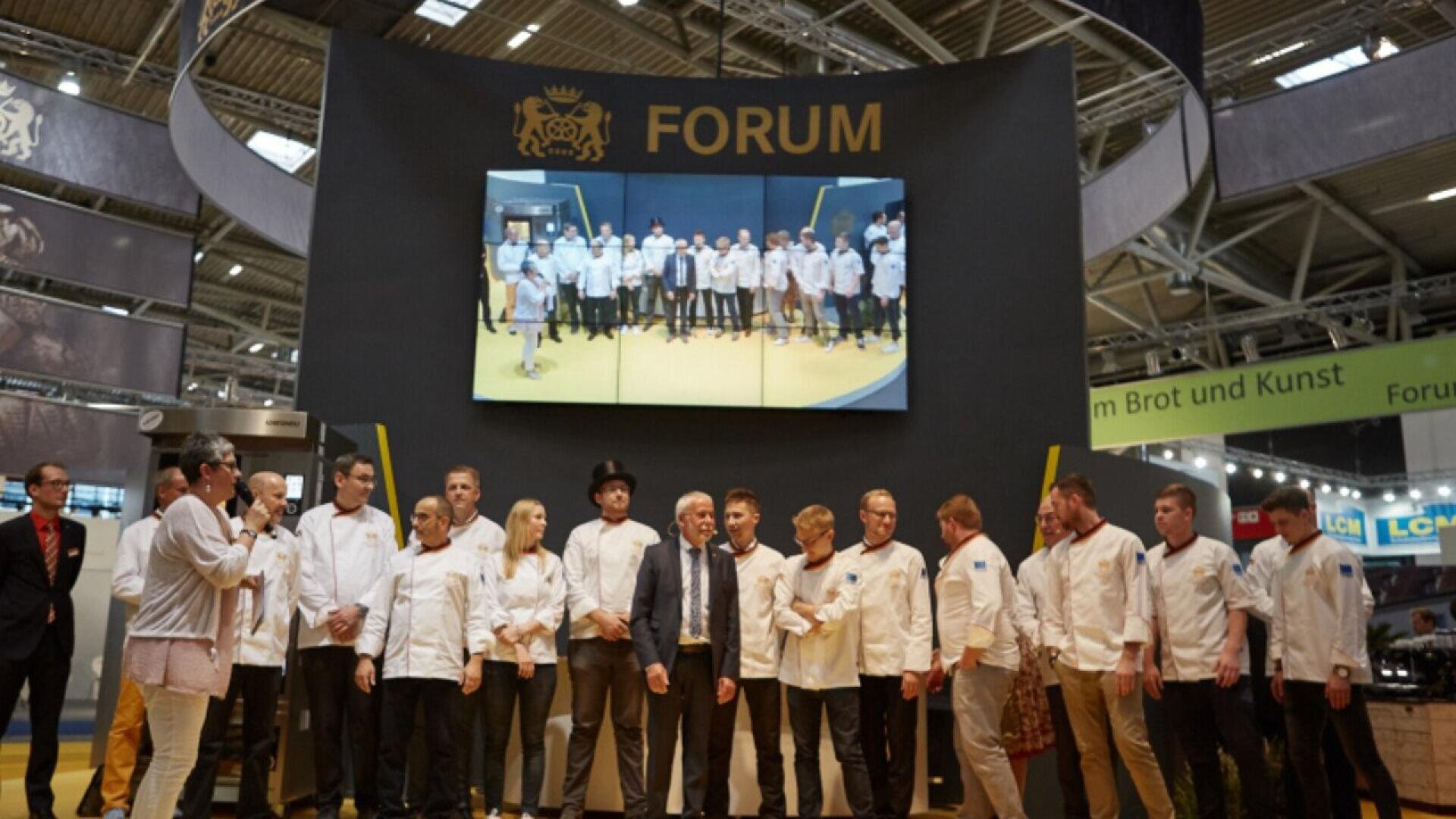 A group of chefs and professionals stand on a stage under a sign that reads FORUM, with a large screen above showing another group photo. The event appears to be held in a spacious exhibition hall.